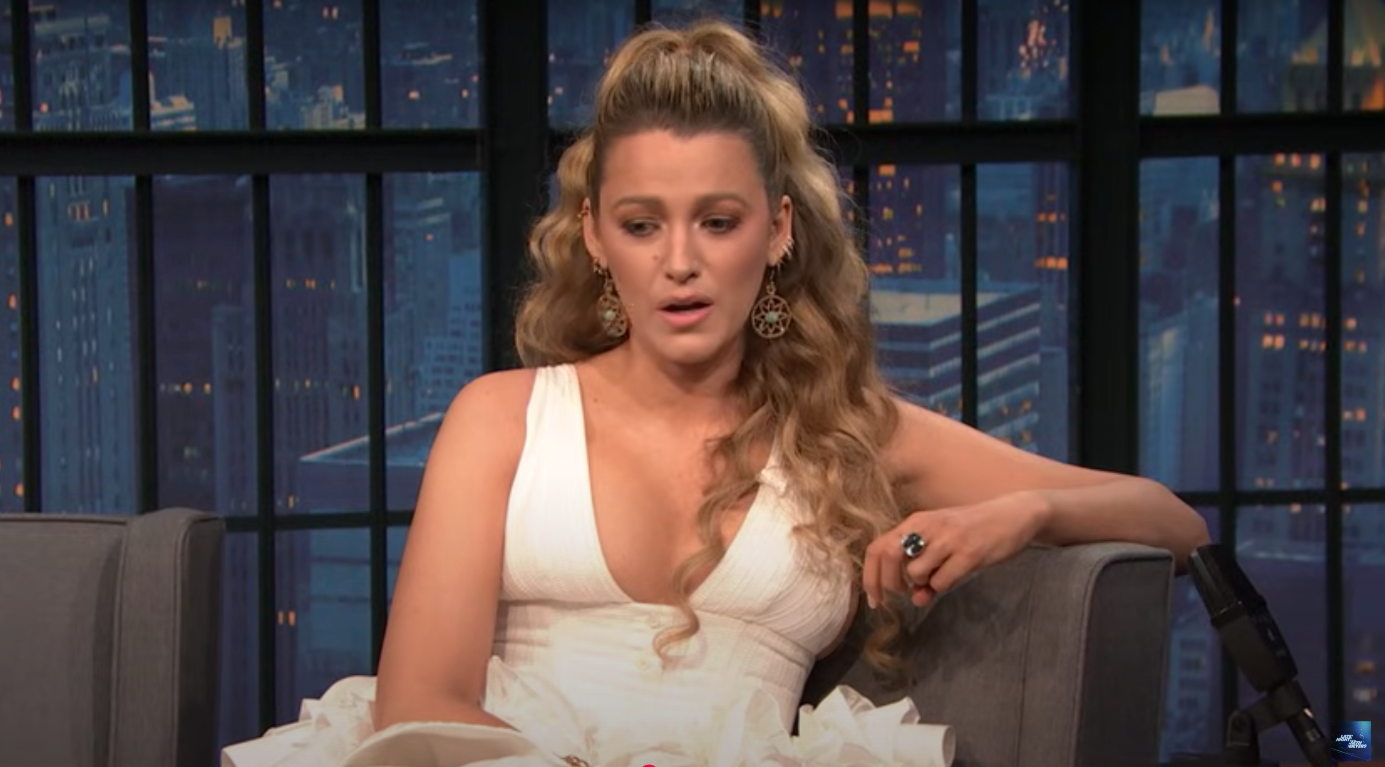Person with long, wavy hair wears a sleeveless dress with a deep neckline, sitting on a talk show set. Cityscape visible in the window behind