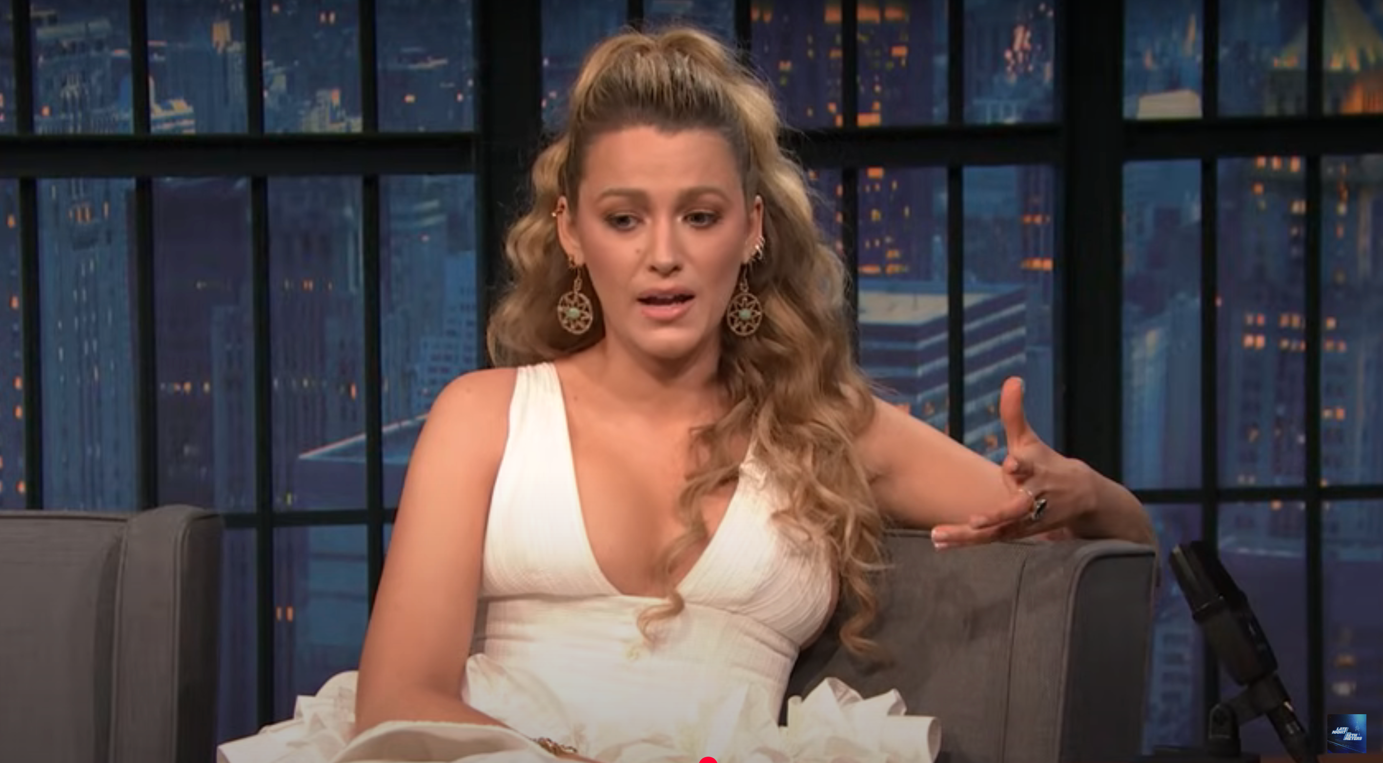 Person with long, wavy hair in a deep V-neck dress gestures while speaking on a talk show set
