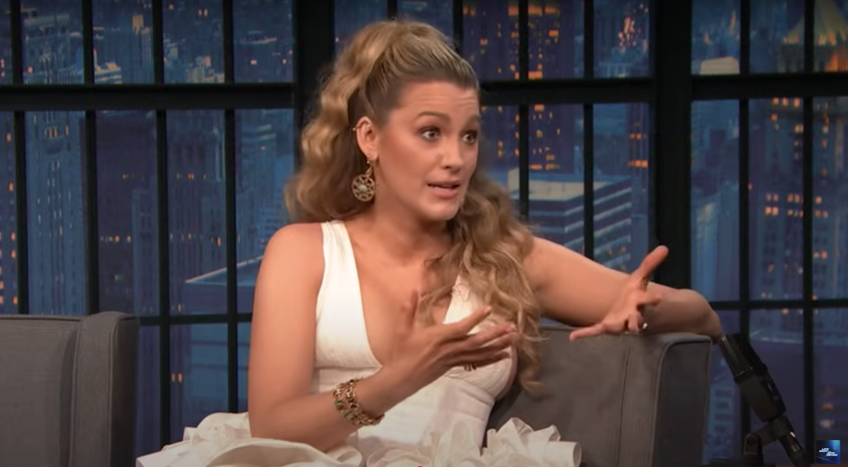 Person with long, wavy hair is sitting and gesturing expressively during a talk show appearance, wearing a sleeveless, ruffled outfit
