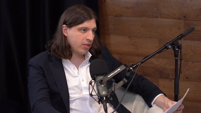 Person in a podcast studio, wearing a blazer and holding papers, speaking into a microphone
