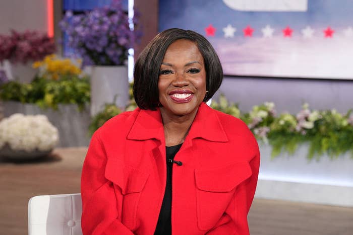 A person in a red coat sits indoors smiling, against a backdrop of flowers and a blurred wall with star decorations