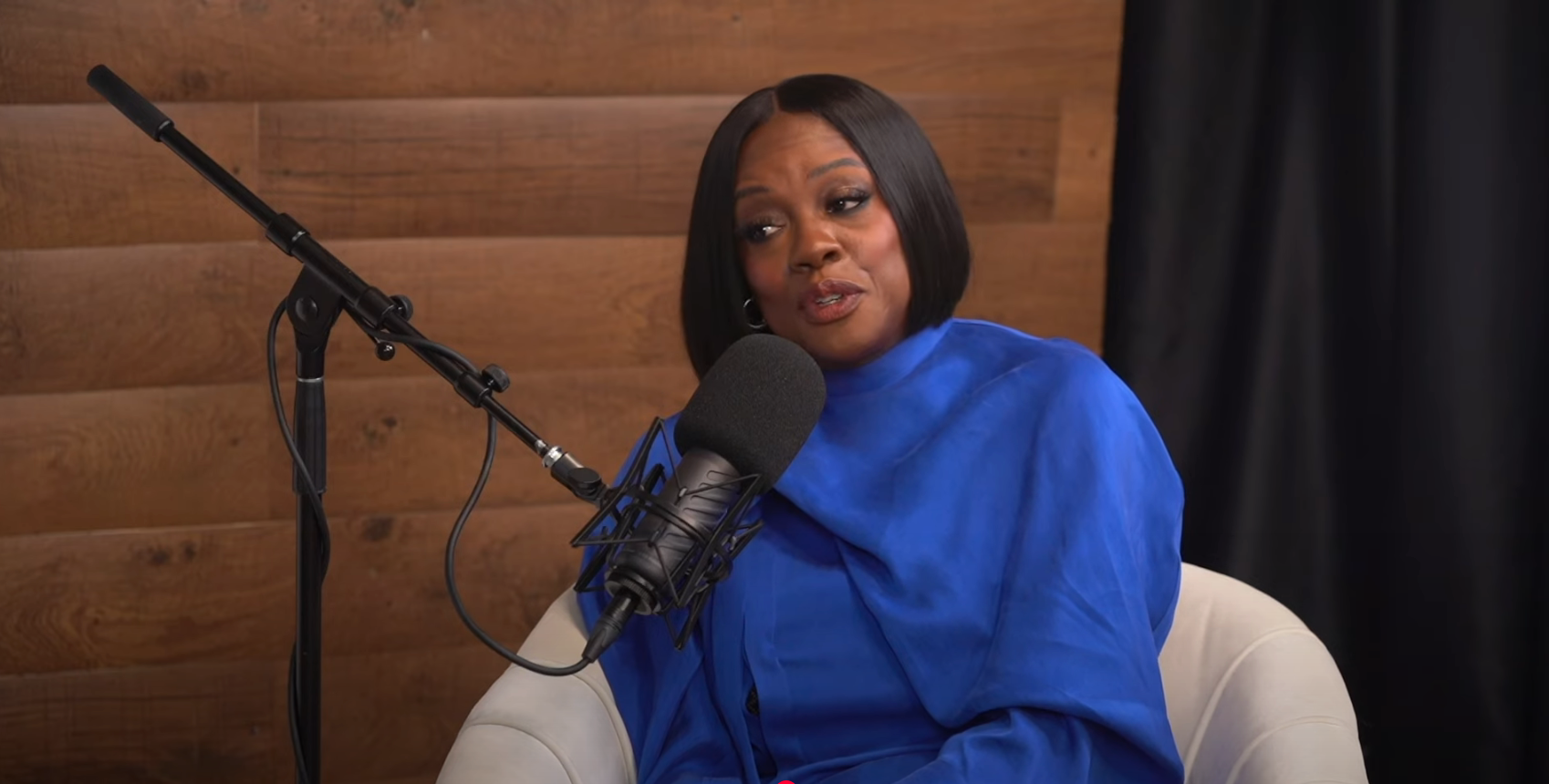 Person in a podcast setting, seated and wearing a stylish outfit with a microphone in front of them