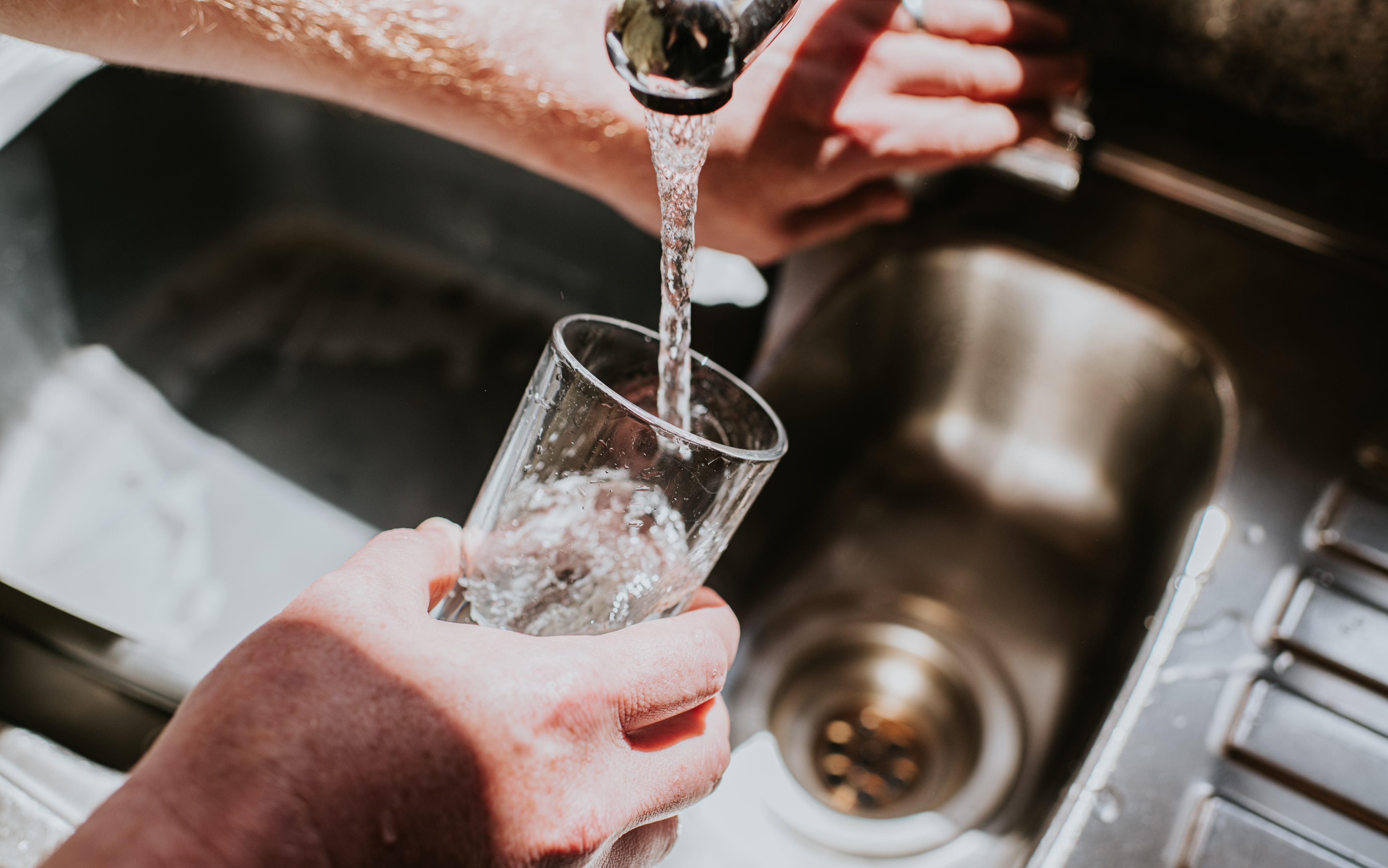 Hands washing a glass under a running faucet in a kitchen sink, illustrating water use or cleanliness