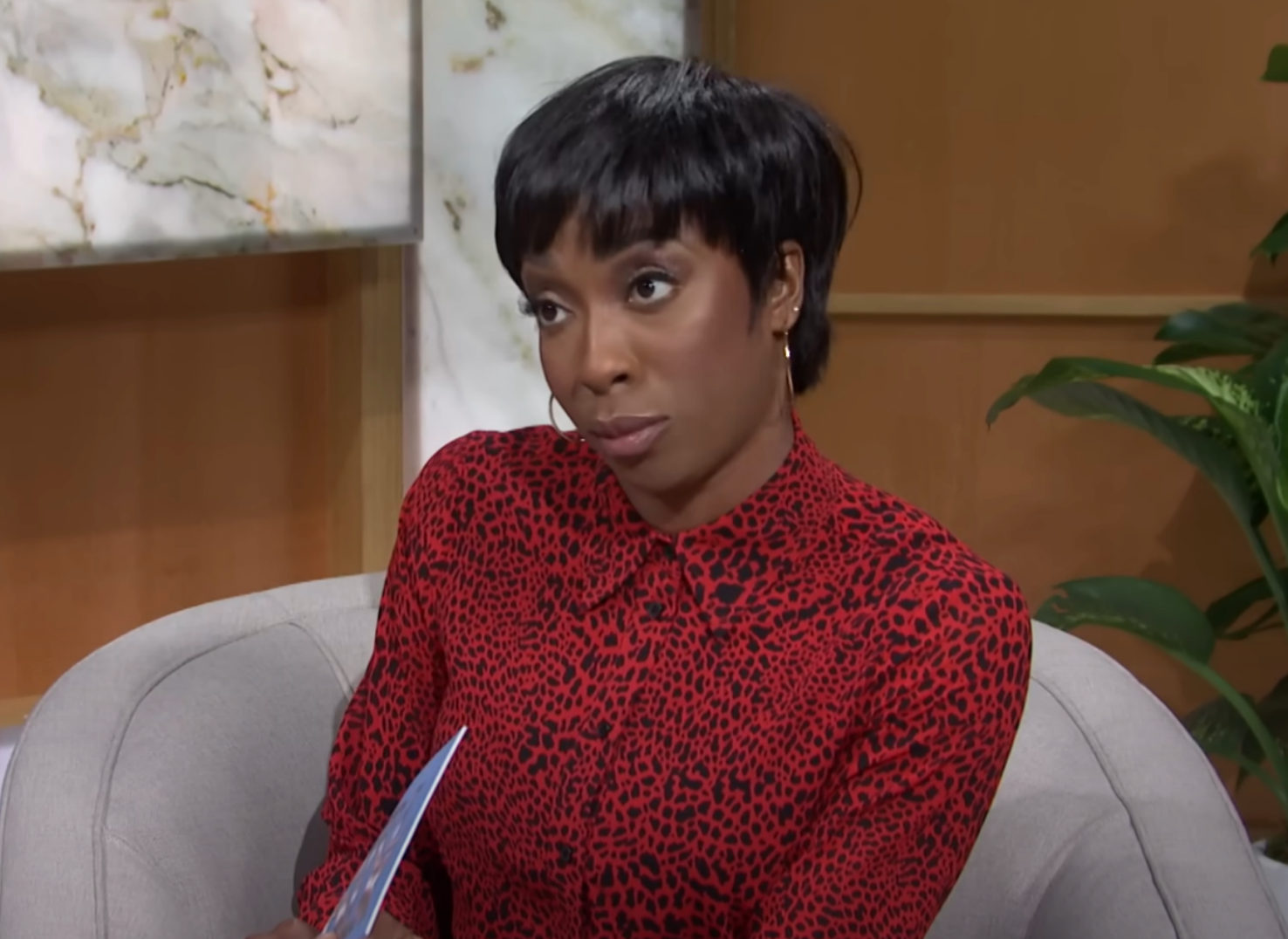 Person sitting indoors, wearing a stylish patterned top, holding papers, with an expressive gaze