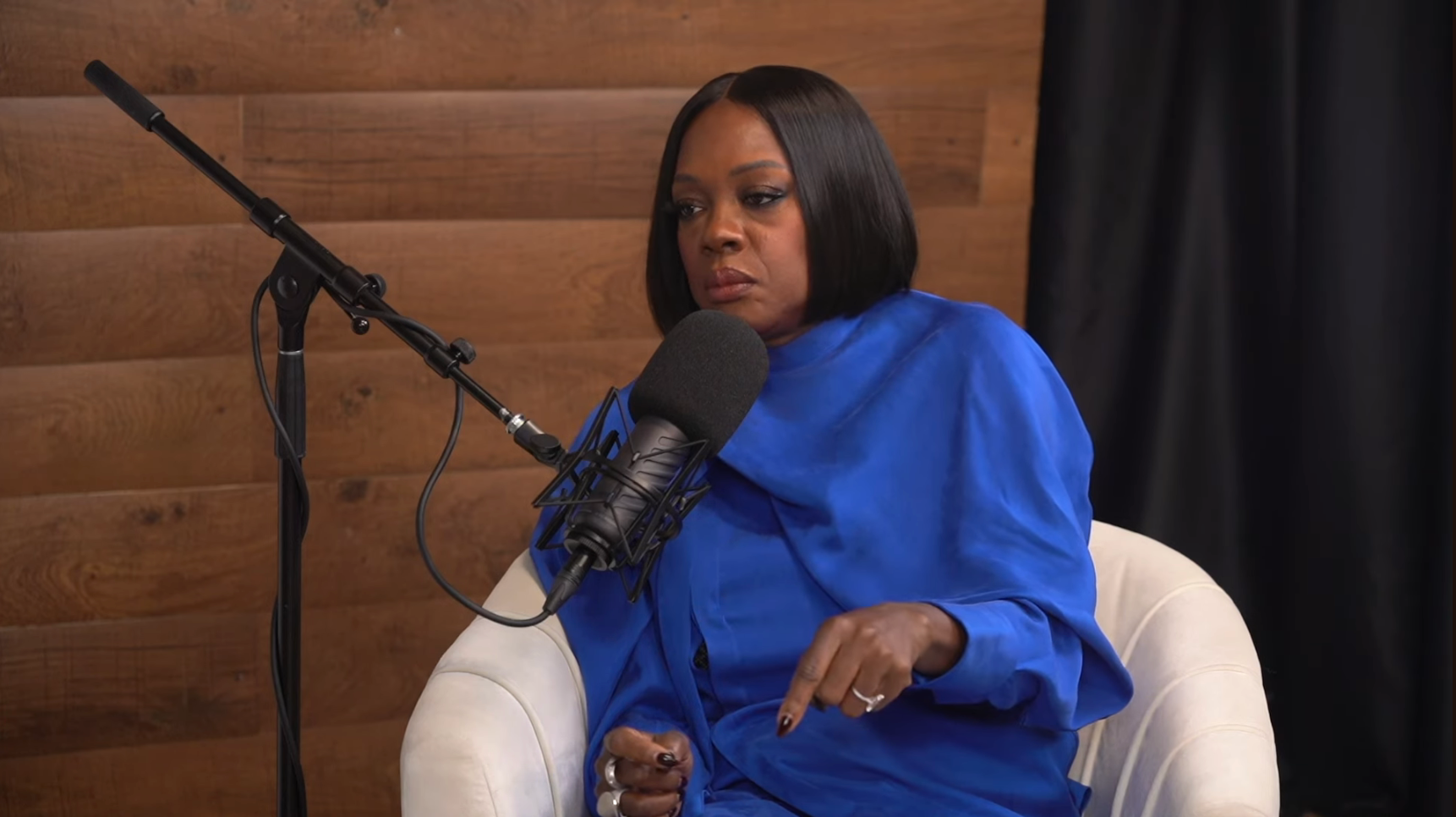 Person in a podcast studio, wearing a sleek outfit, speaking into a microphone while seated in a white chair