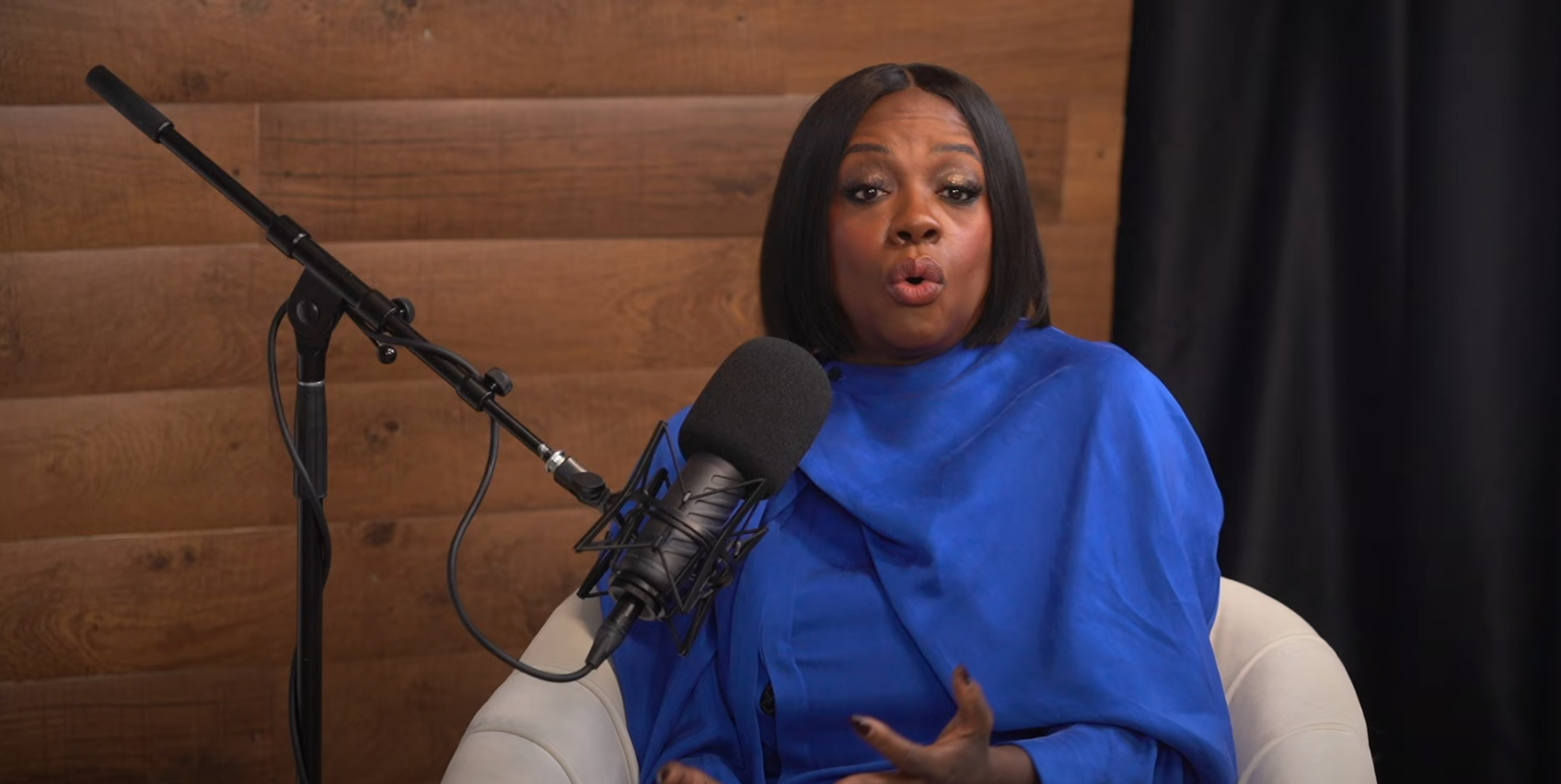 Person in a podcast studio, wearing a stylish draped outfit, speaking animatedly into a microphone