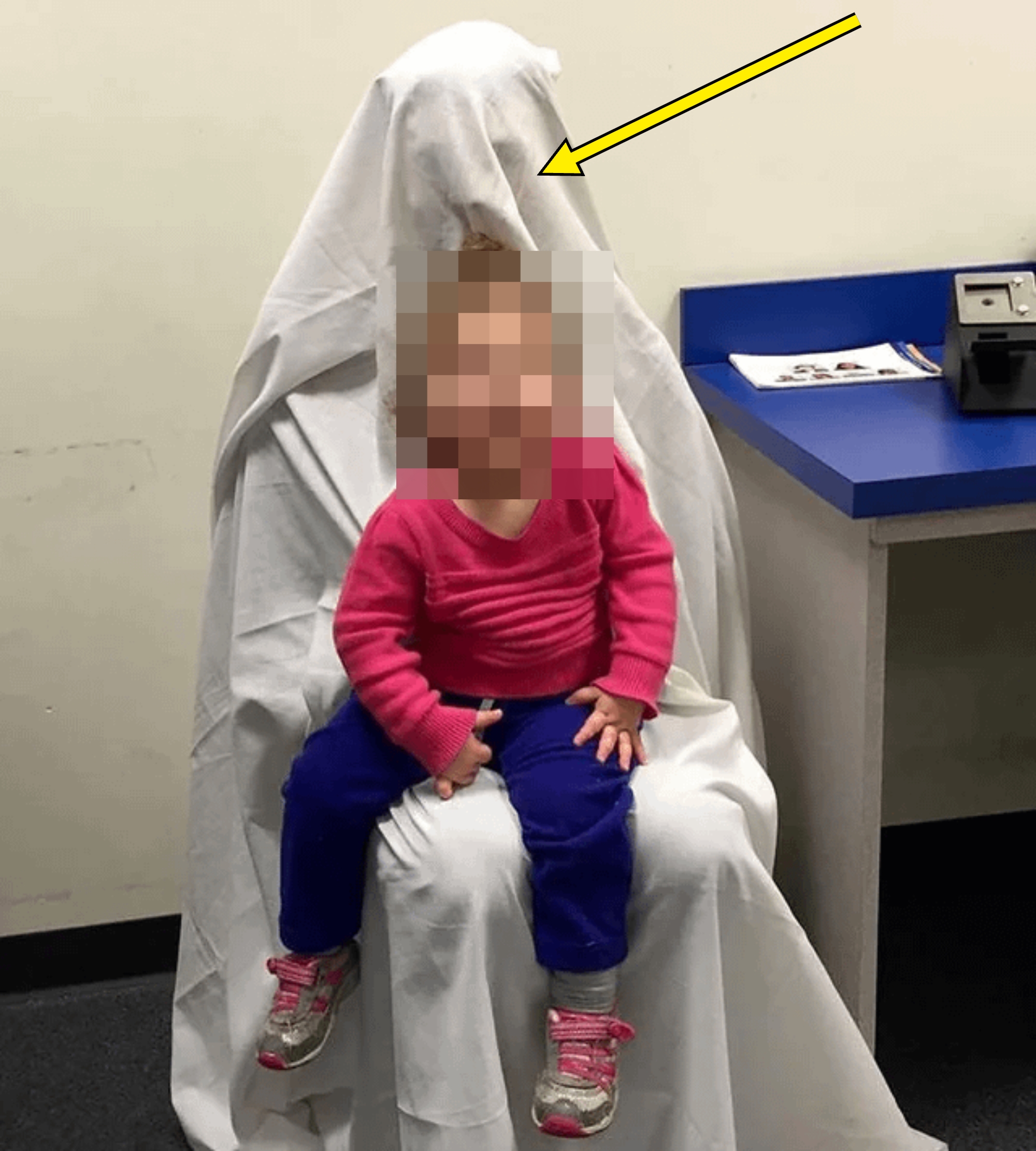 Child sits on an adult draped in a sheet, resembling a ghost, in an office setting