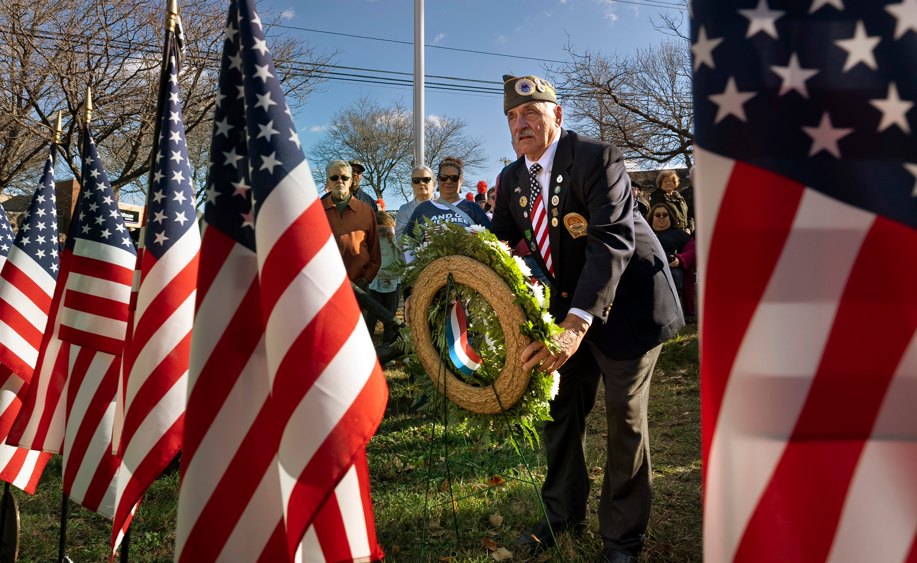 A veteran wearing a military cap places a wreath at a ceremony, surrounded by American flags and onlookers