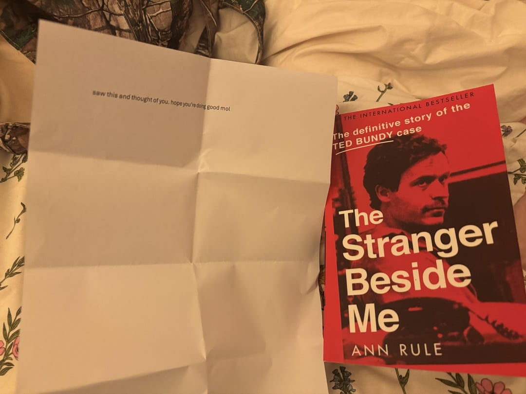 A folded note with a message and a book titled "The Stranger Beside Me" by Ann Rule are shown lying on a bed