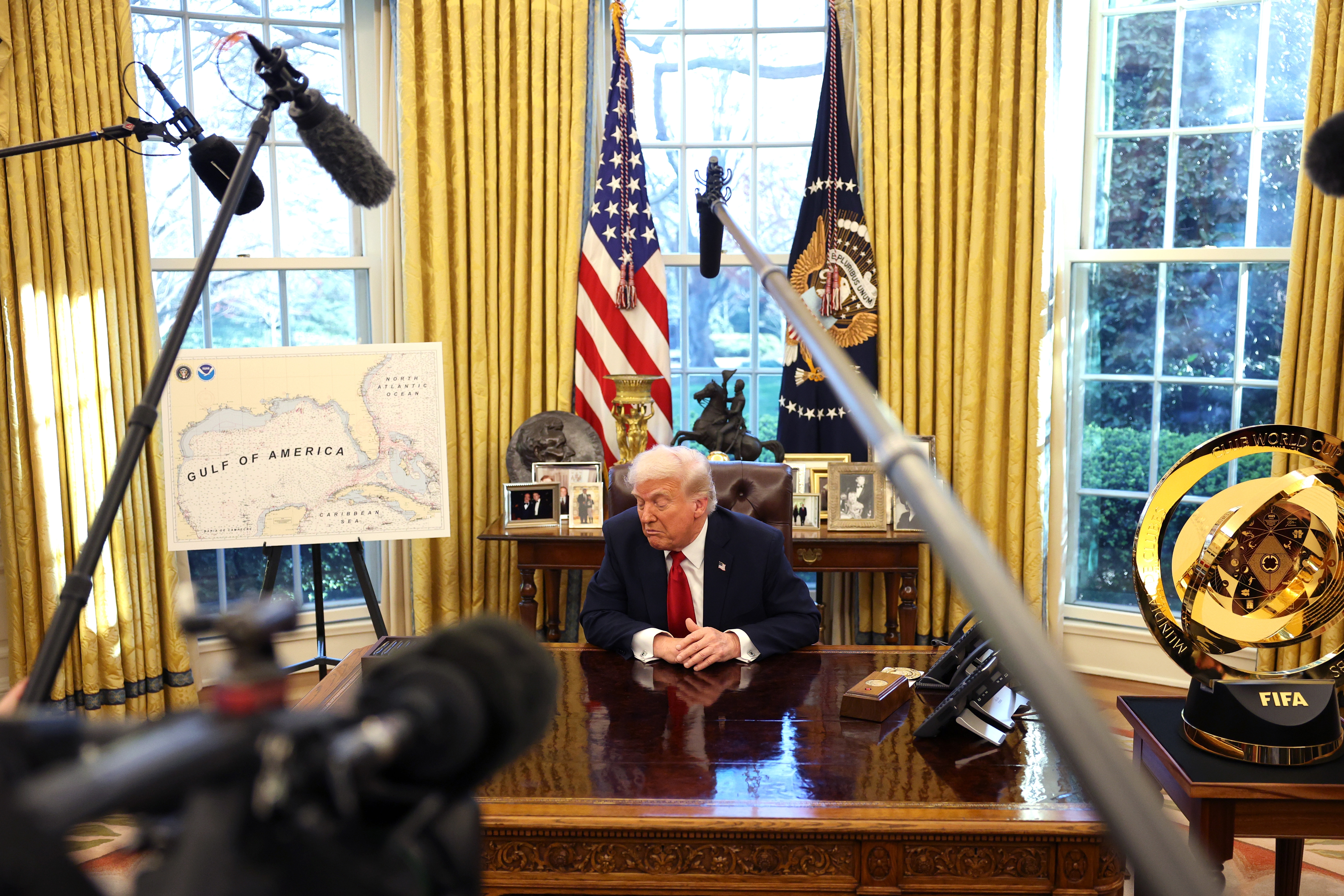 A person seated at the Oval Office desk, surrounded by microphones. A map and a trophy are visible nearby