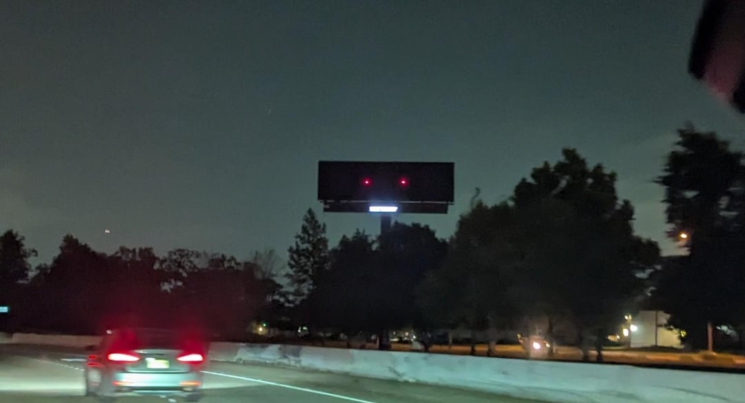 A dark, blank billboard with two small red lights, seen from a road surrounded by trees at night