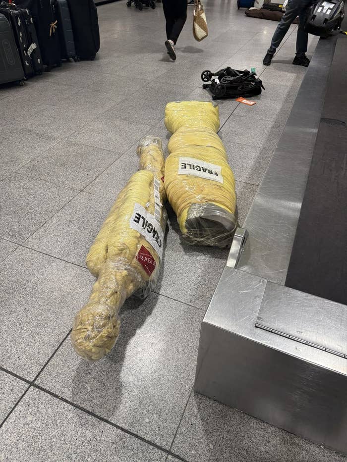 Two large, yellow, wrapped items labeled "FRAGILE" and "HANDLE WITH CARE" lie near an airport baggage carousel
