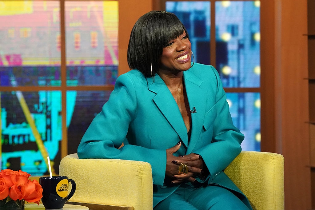 Person smiling in a bright suit while sitting on a talk show set, holding a coffee mug