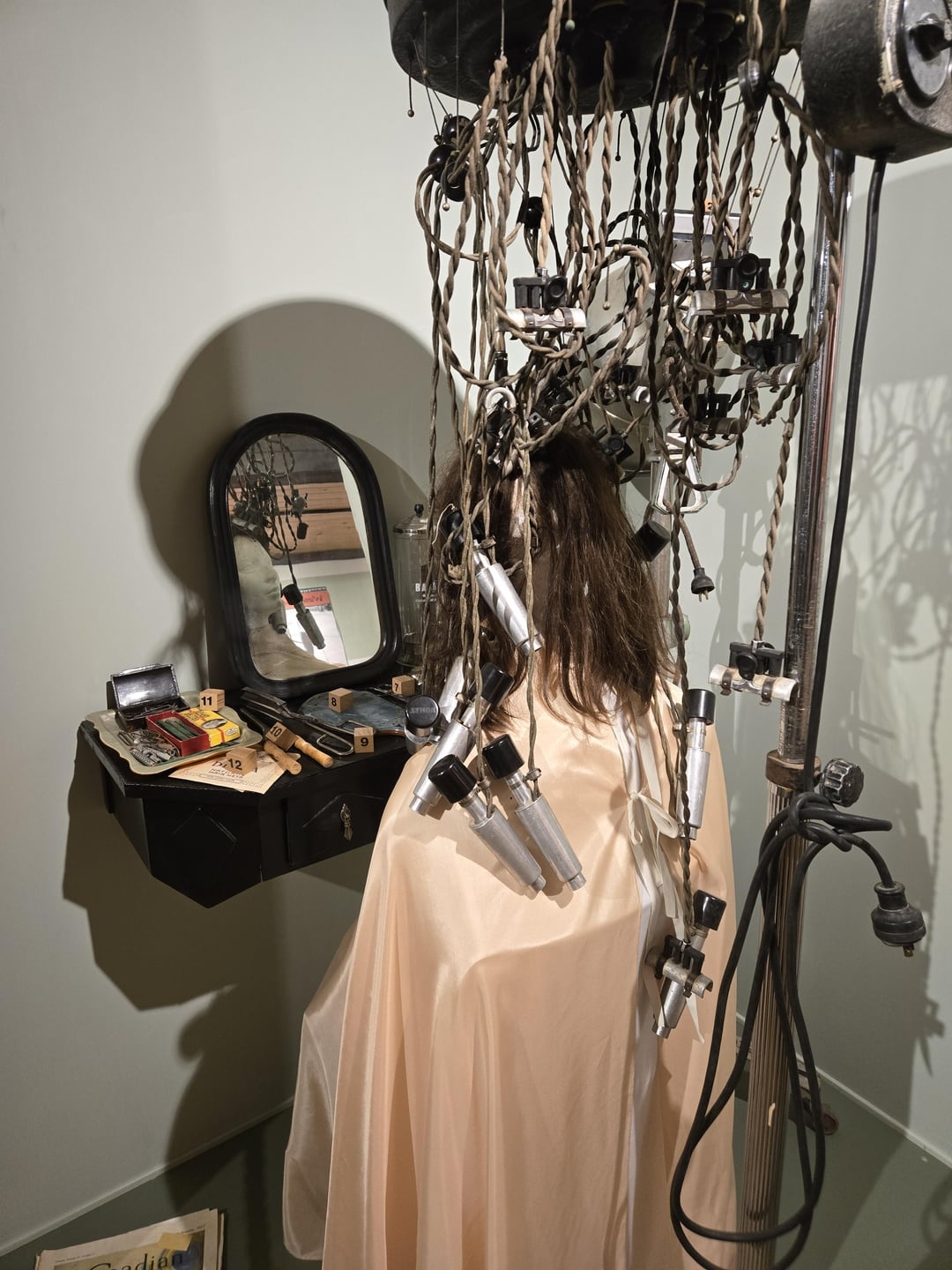 A mannequin under a vintage hair salon dryer with tangled wires, sitting by a vanity with old grooming tools