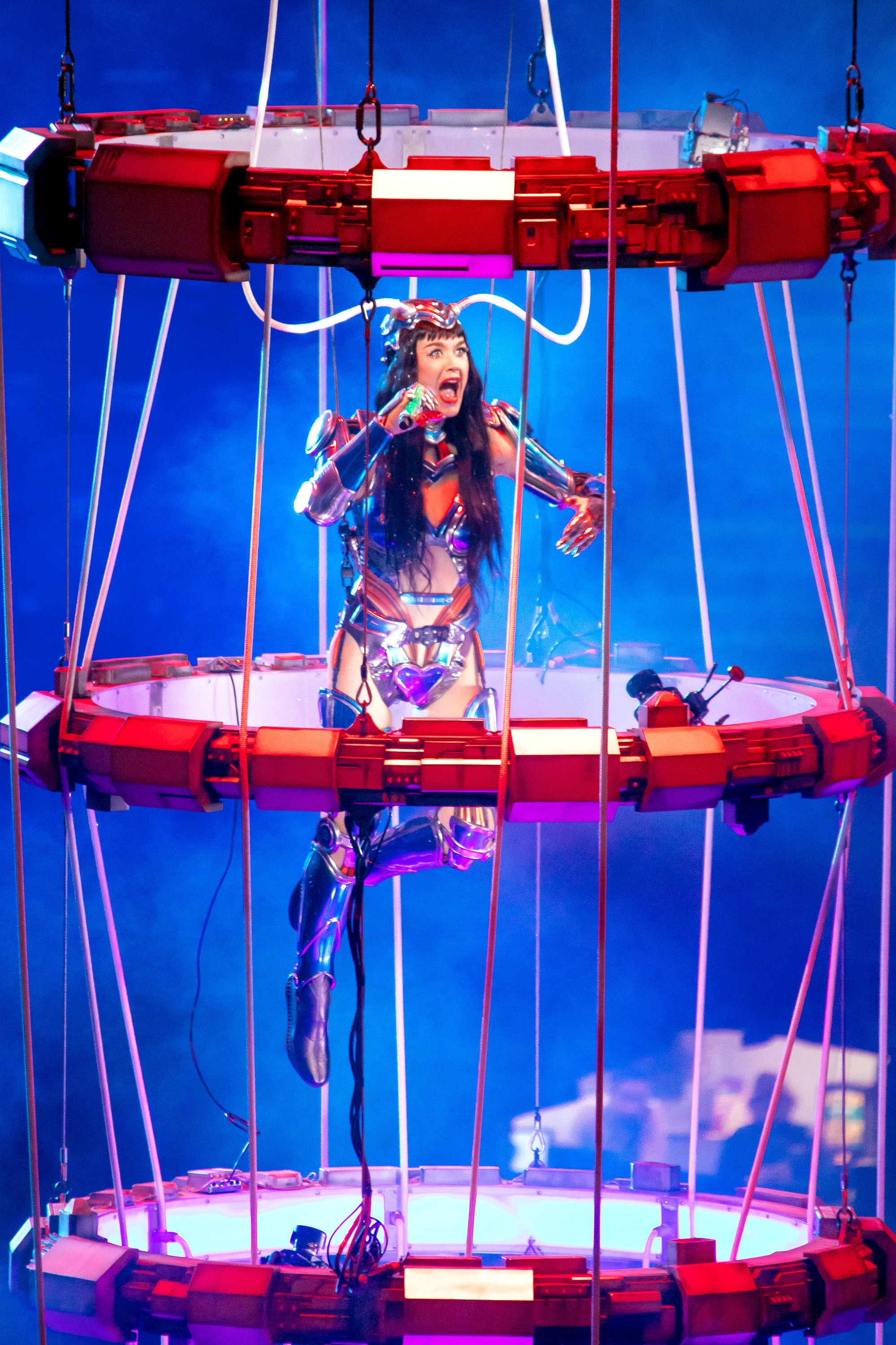 Performer in futuristic costume sings energetically while suspended in a circular stage apparatus