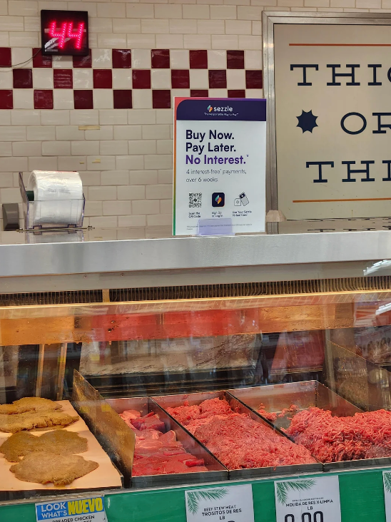 Butcher counter with various meats displayed. A sign above promotes a "Buy Now, Pay Later" plan with four interest-free payments