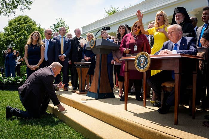 Group of people in a garden setting; one man signs documents at a table with presidential seals, while another kneels on the grass nearby