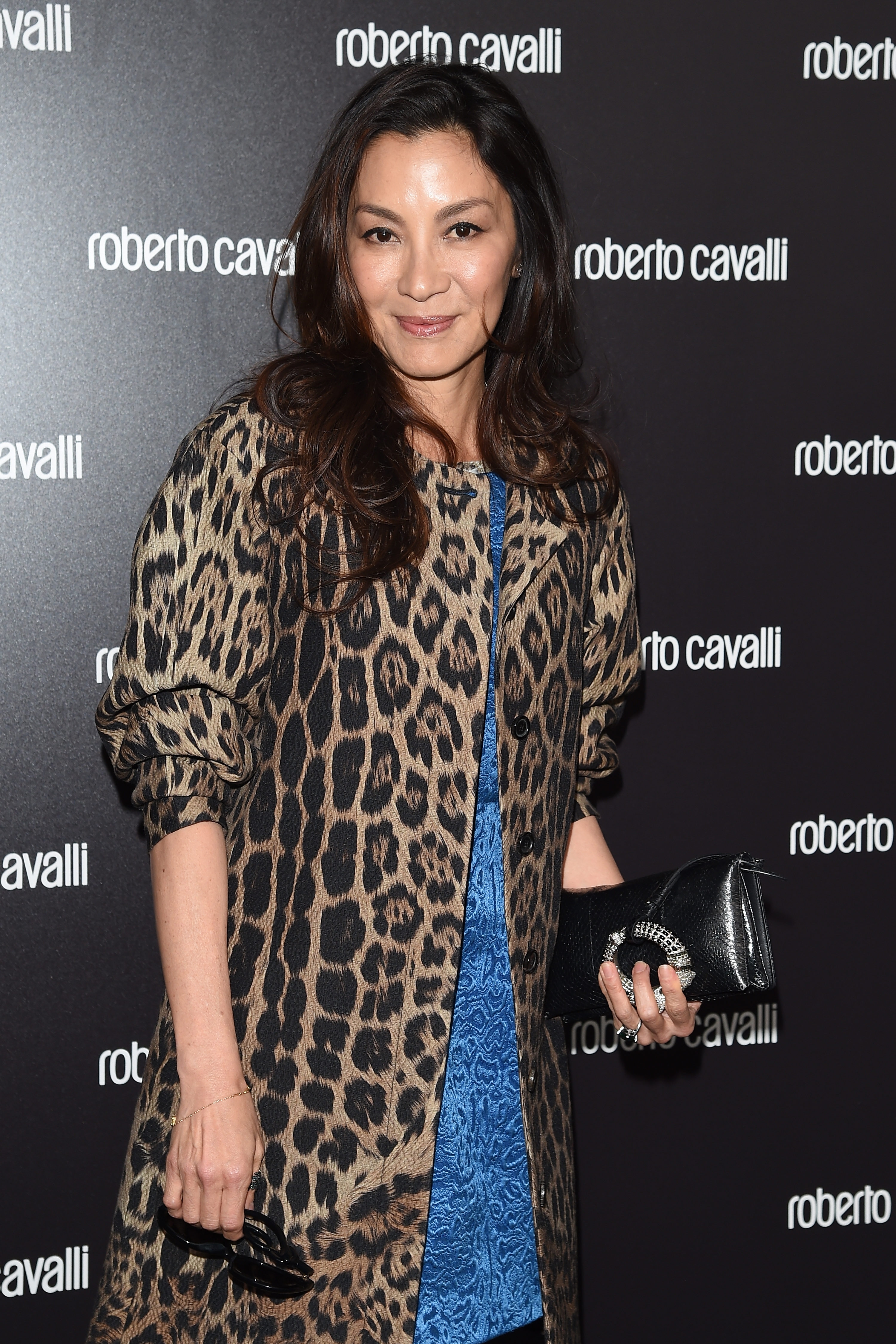 Person wearing a leopard print coat over a textured blue dress, holding a black clutch at a Roberto Cavalli event