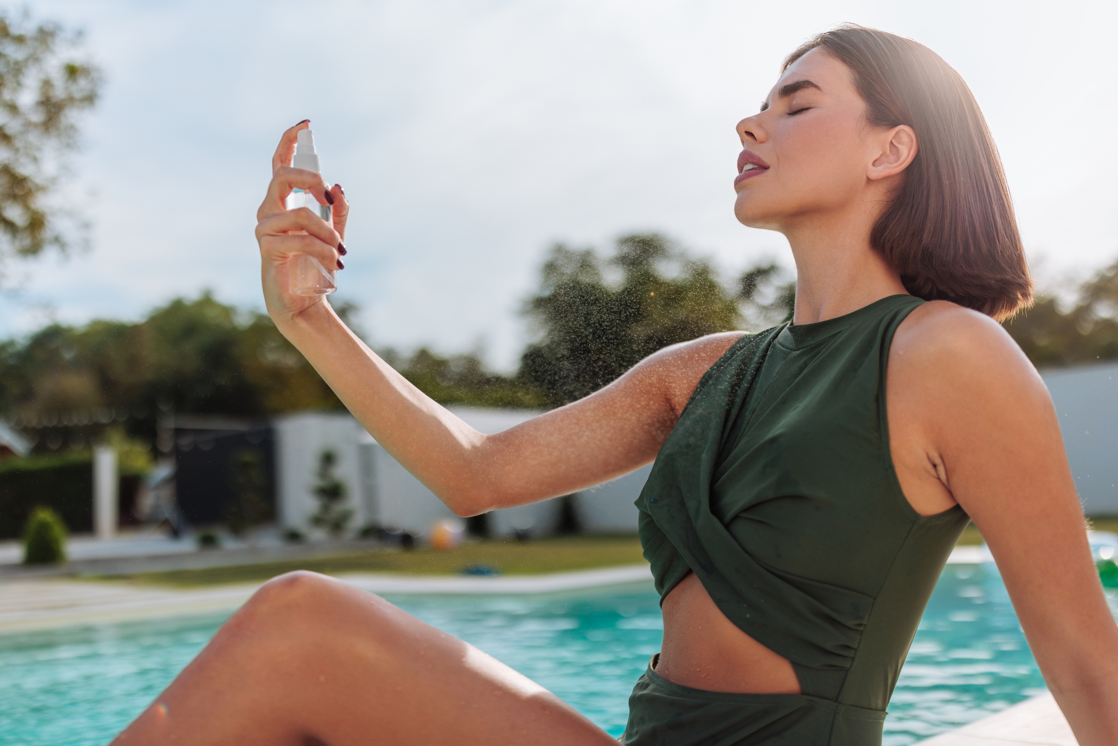 Person by a pool, wearing a stylish sleeveless cutout dress, spritzing face with mist, eyes closed, enjoying a moment of relaxation outdoors