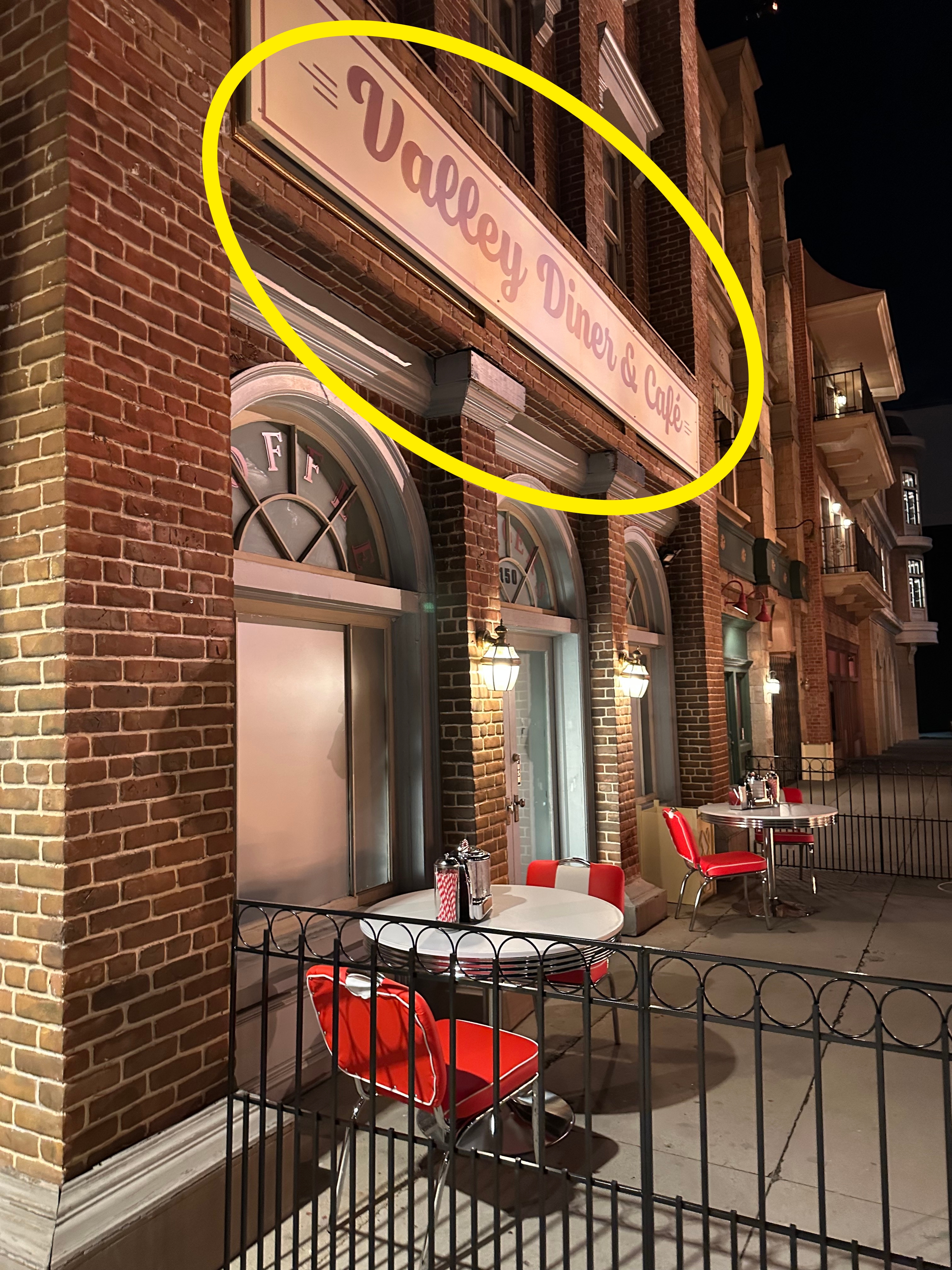 Night view of a vintage diner with outdoor seating, rounded tables, and red chairs, set against a backdrop of brick buildings