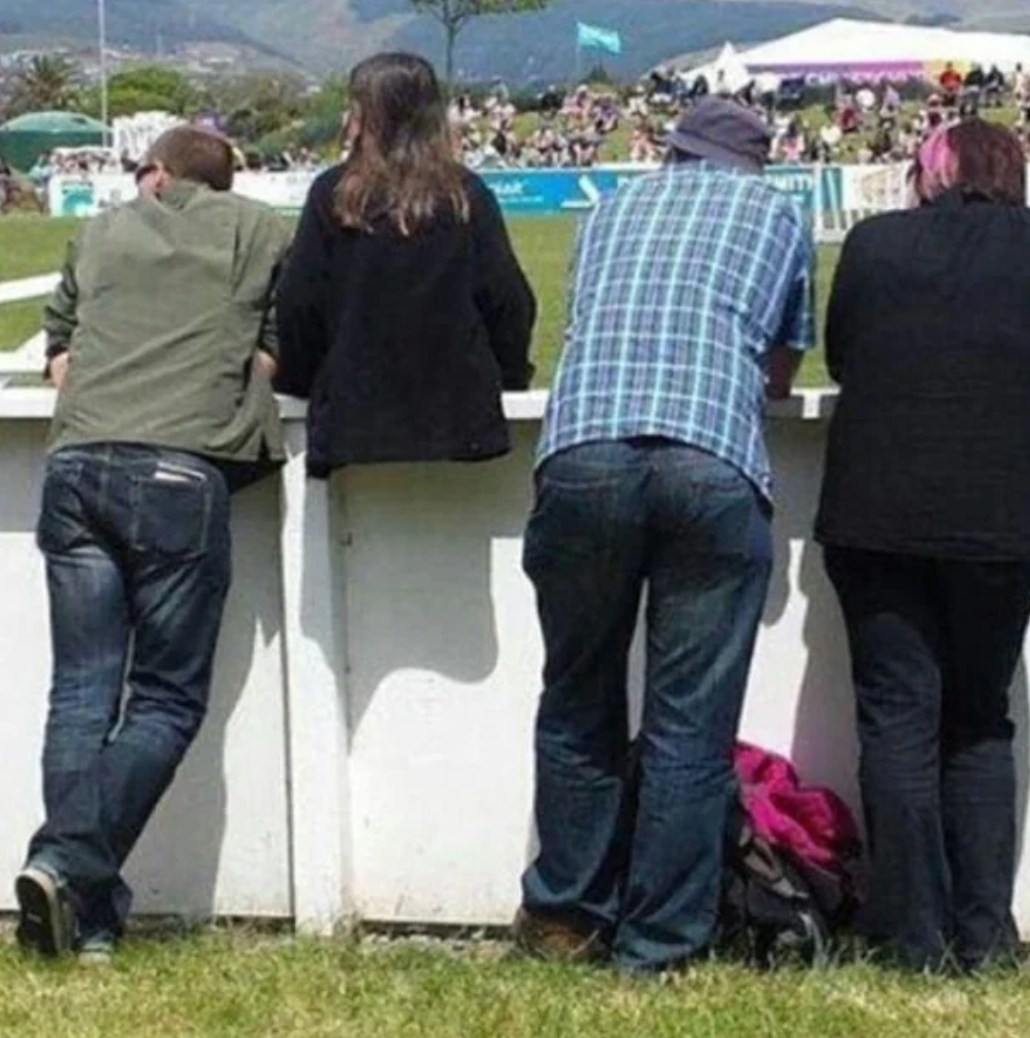Four people leaning over a fence, viewing an outdoor event. Casual attire