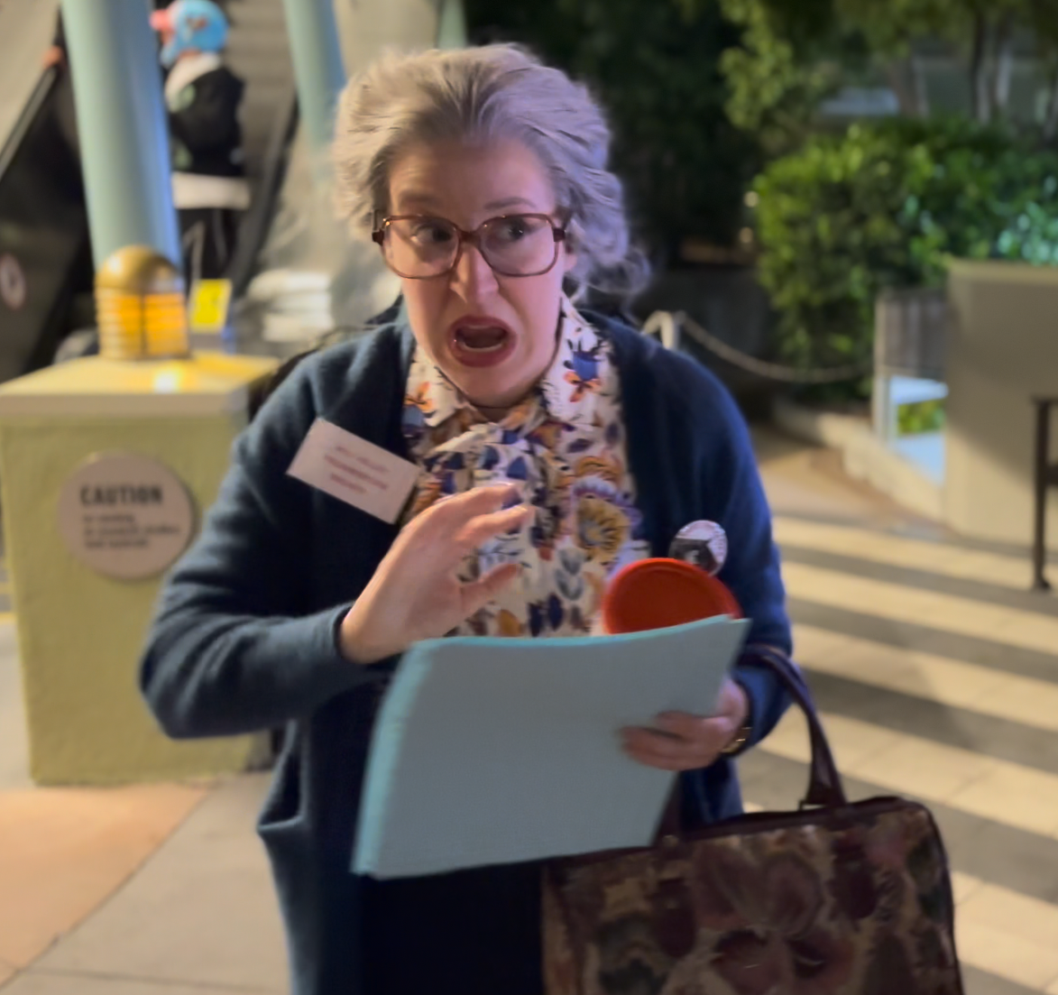 Person in patterned blouse and cardigan, holding a binder and mug, looks surprised. Background includes part of a street scene