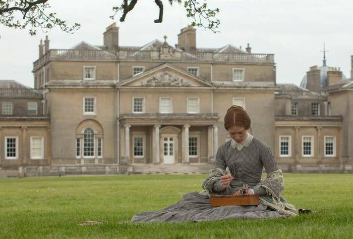 Persona vestida en estilo del siglo XIX, sentada en un césped, borda frente a una gran mansión de estilo clásico en el fondo