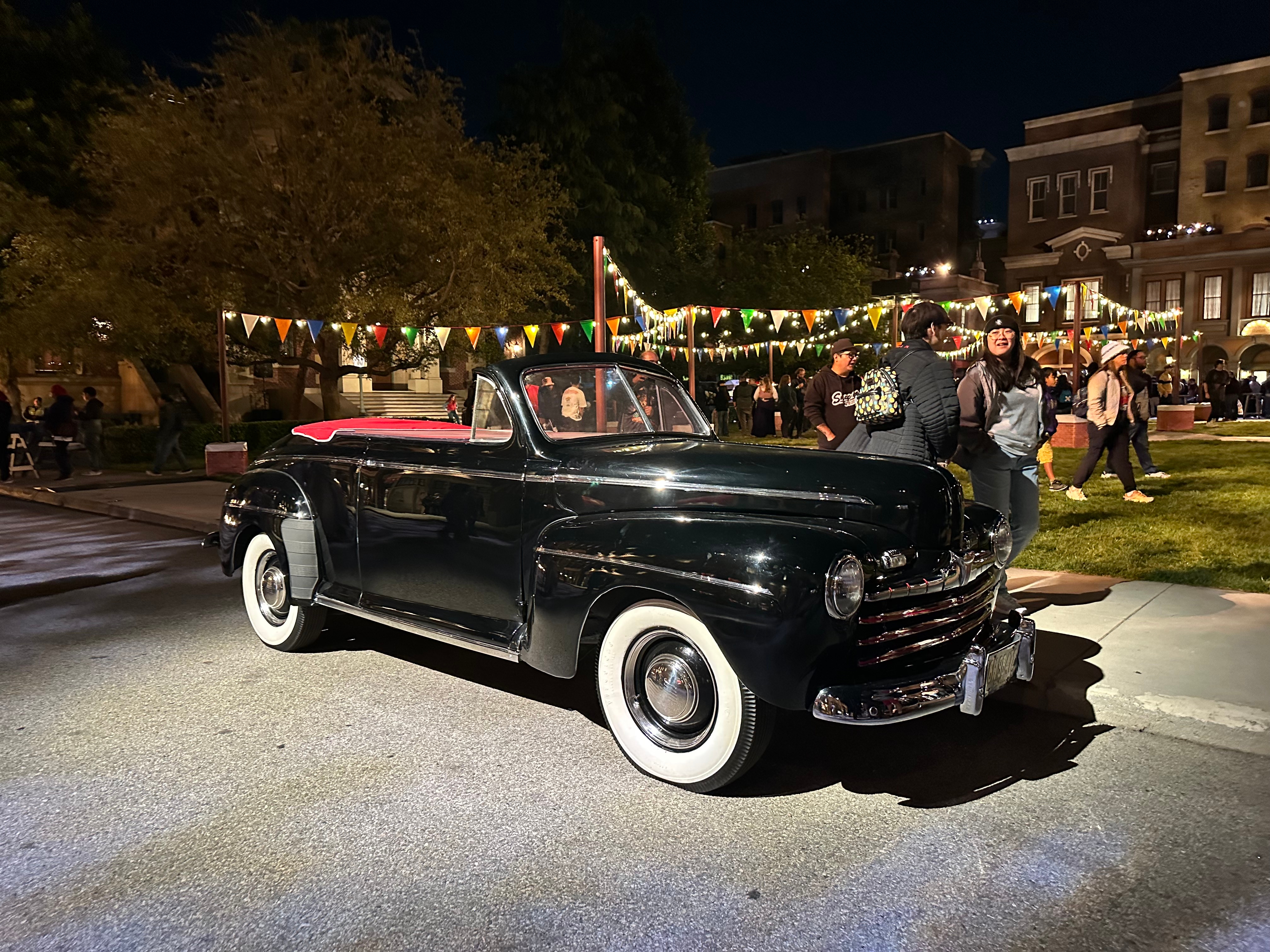 Vintage convertible car parked at night during an outdoor event with people in the background under string lights and colorful pennants