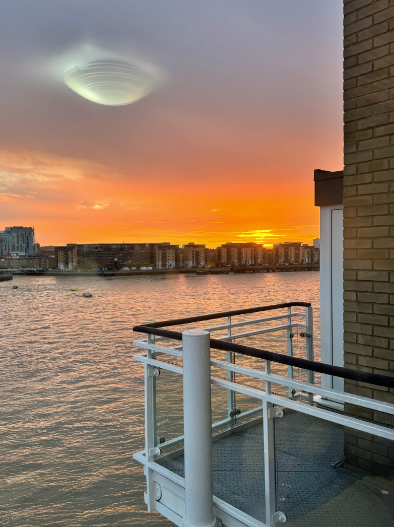 Sunset view over a city river from a balcony, with a distant glowing disk-shaped object in the sky, resembling a UFO