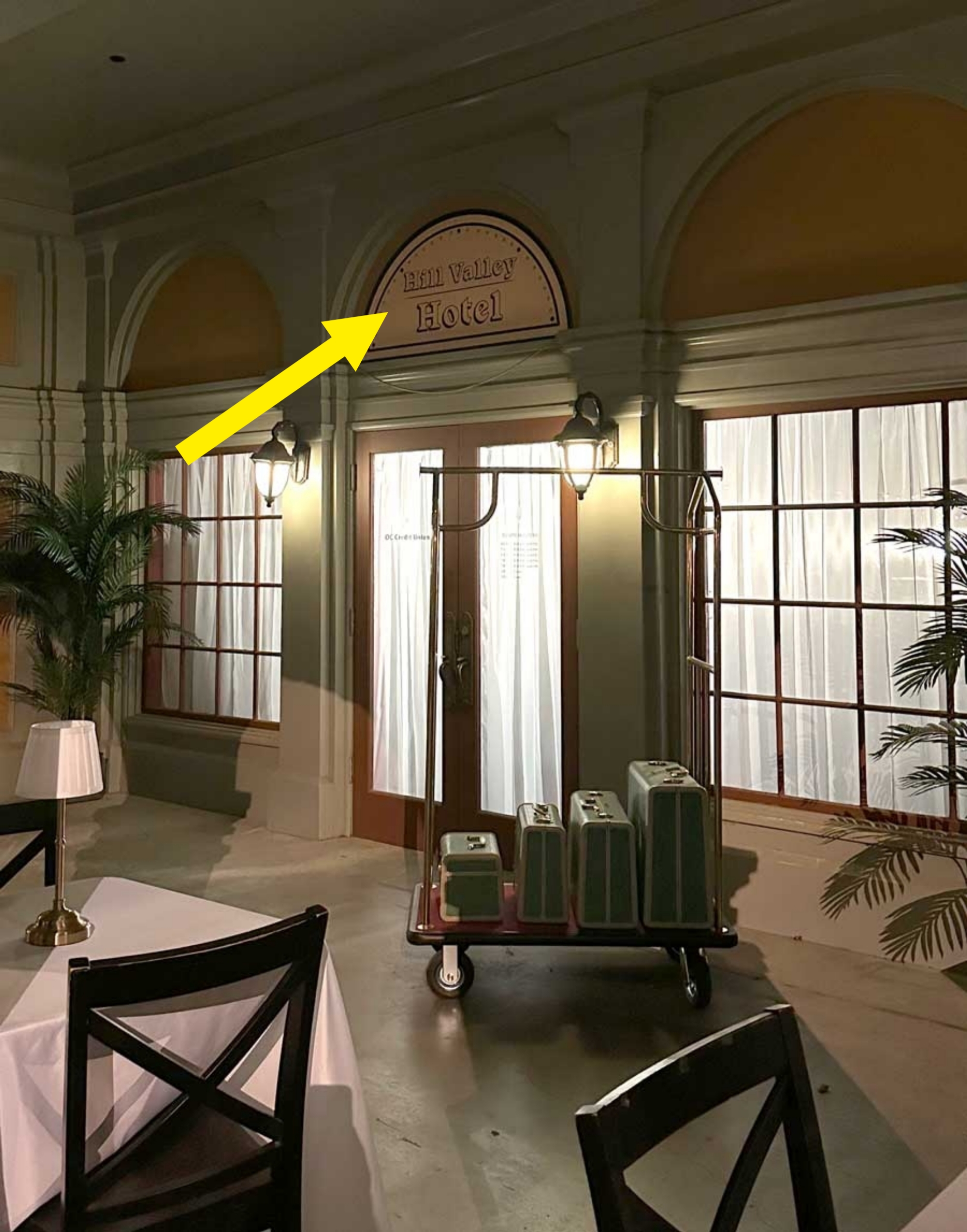 Hotel façade at night with a luggage cart holding suitcases outside and a dining table in the foreground