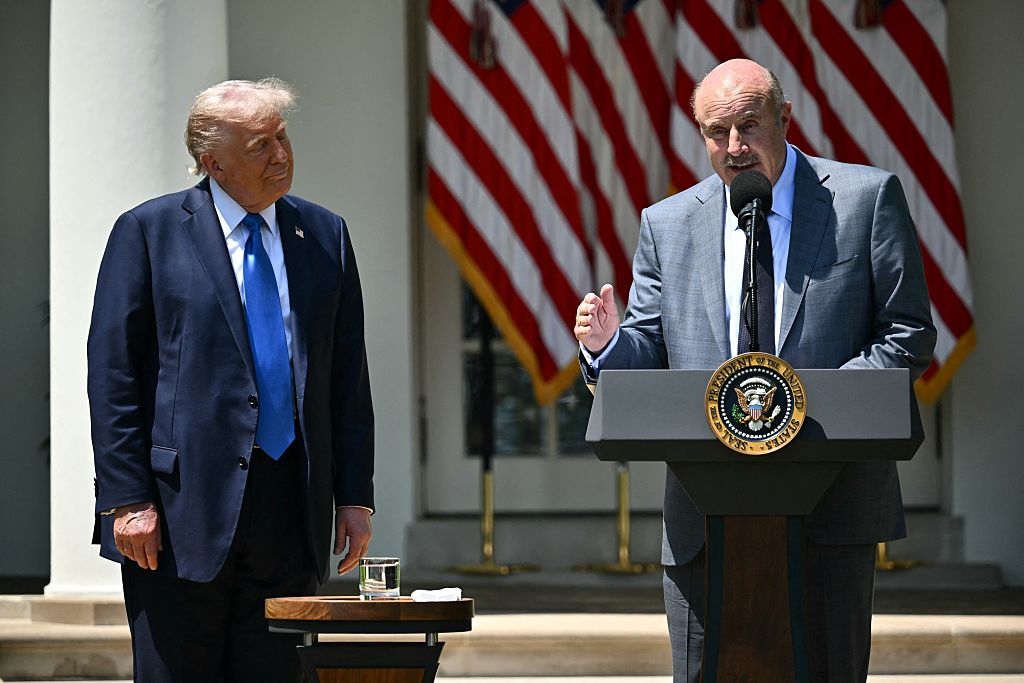 Two individuals stand at podiums. One speaks into a microphone; the other listens, standing beside an American flag backdrop