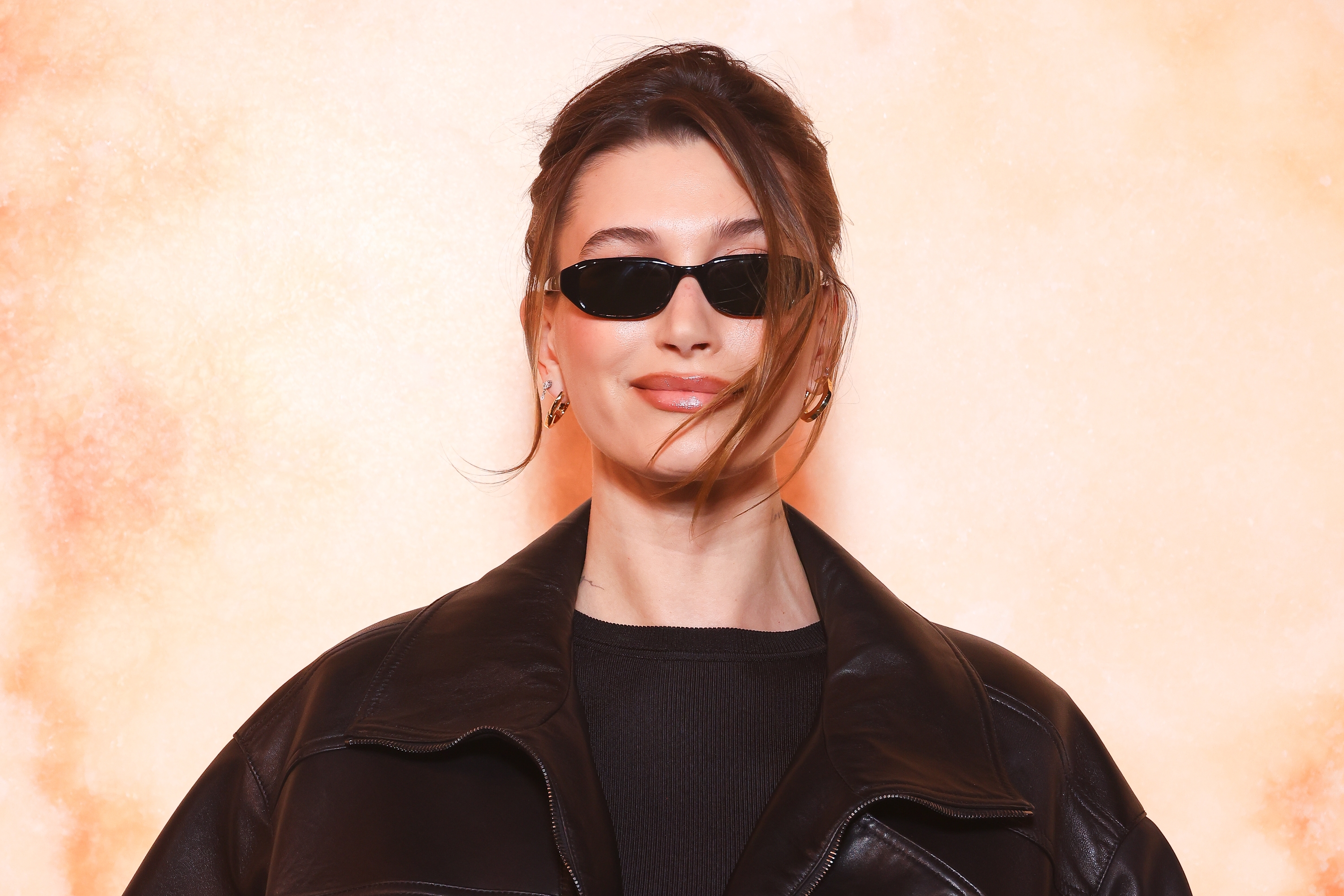 Person in a black leather jacket and sleek sunglasses, smiling at the camera. Hair is styled up with loose strands framing the face