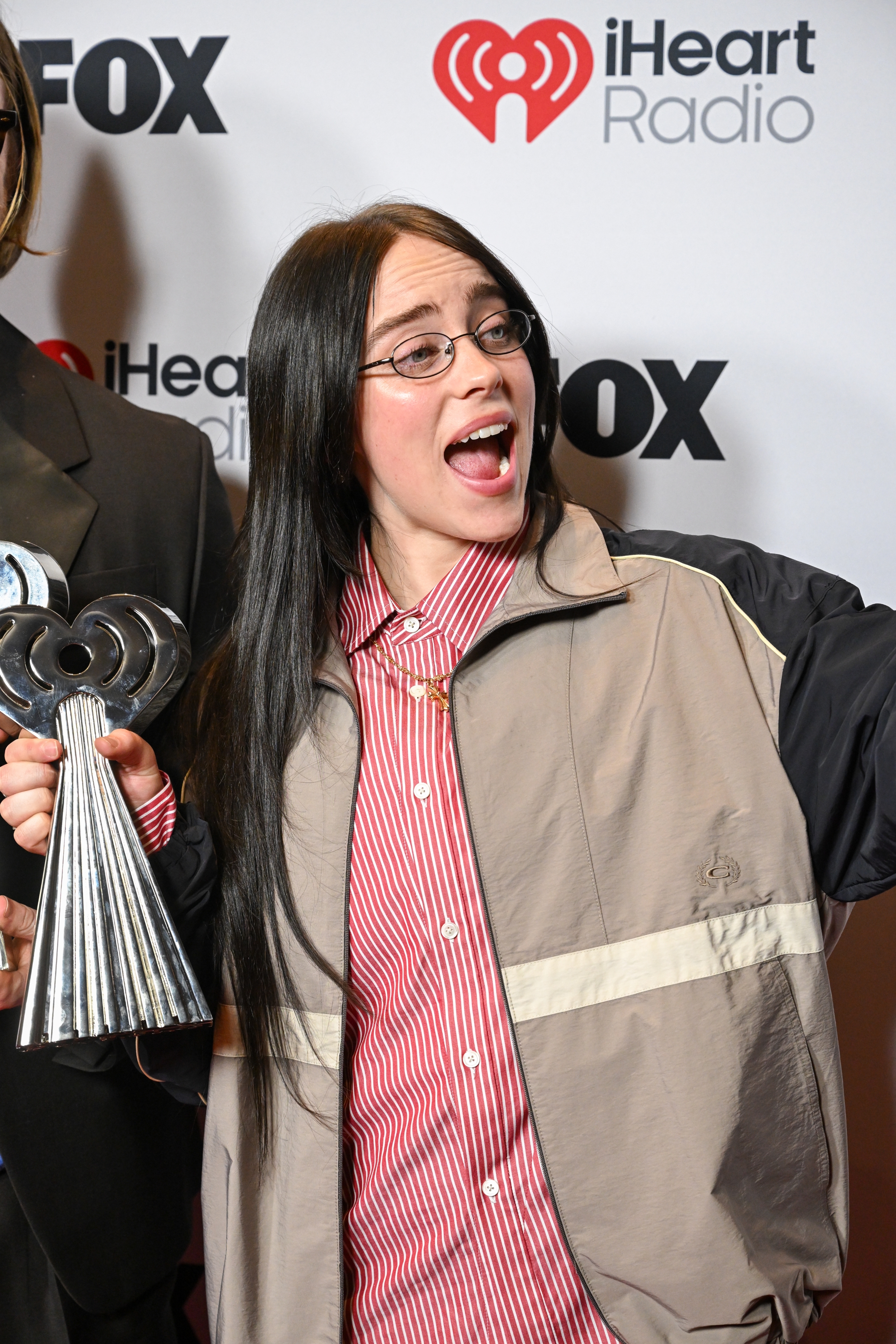 Individual on a red carpet holding an award, wearing a striped shirt and jacket, with long dark hair and glasses, appearing joyful