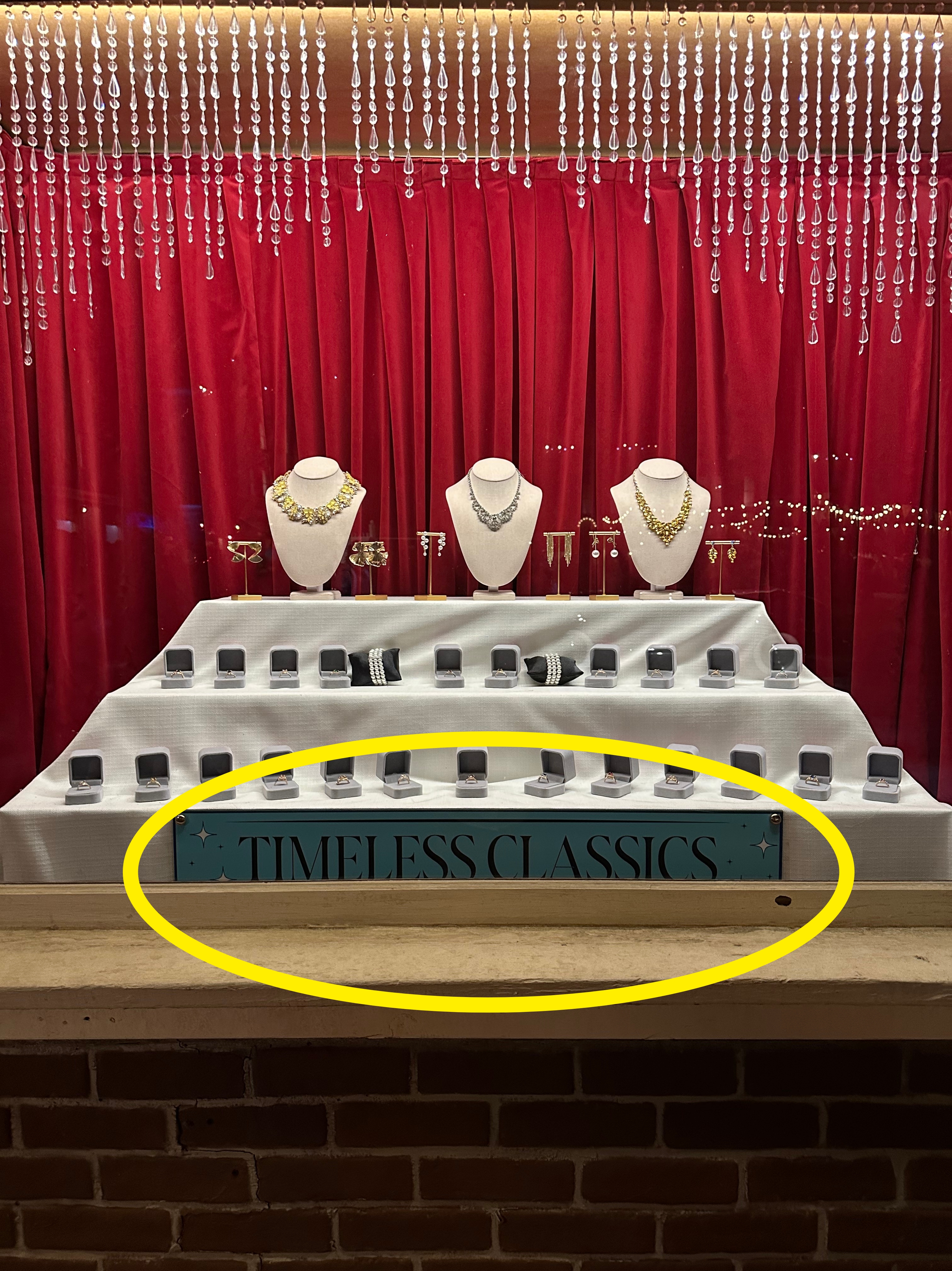 Jewelry store window with necklaces on stands and rows of rings, displayed against a curtain backdrop. Sign reads &quot;Timeless Classics.&quot;