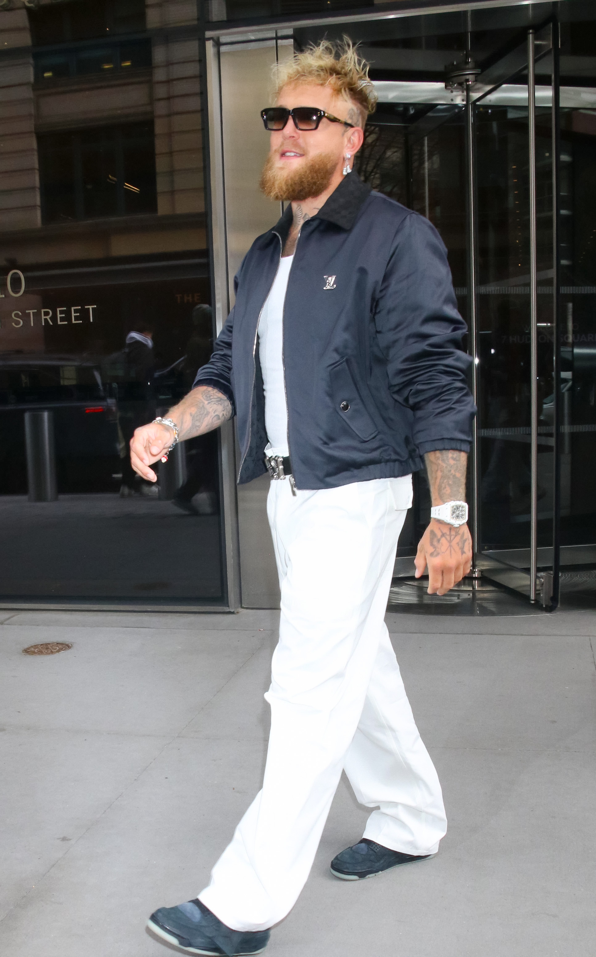 Person wearing a casual jacket, white shirt, and loose white pants with sunglasses and wristwatch, walking confidently outdoors