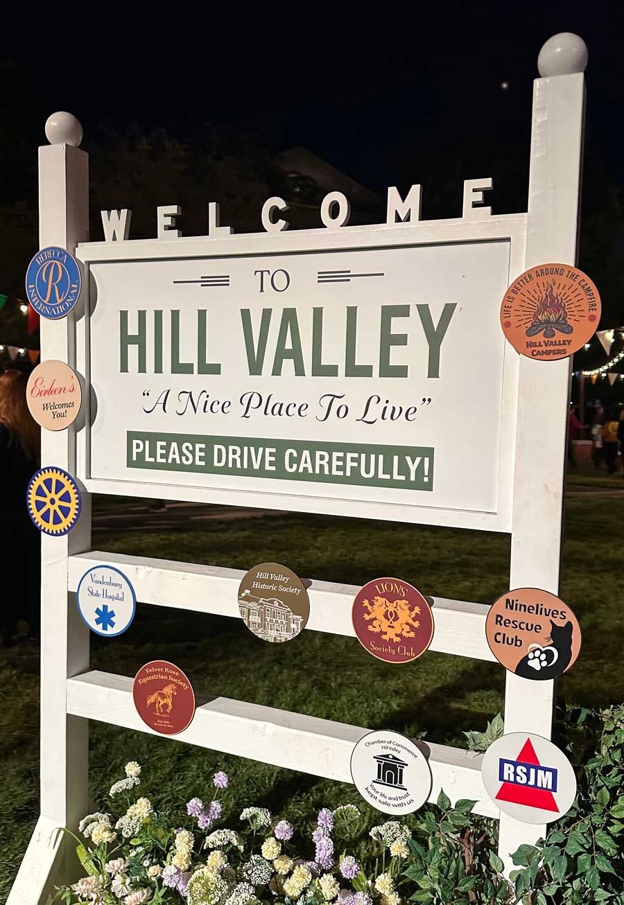 Wooden sign reading &quot;Welcome to Hill Valley: A Nice Place to Live. Please Drive Carefully!&quot; with various club emblems attached