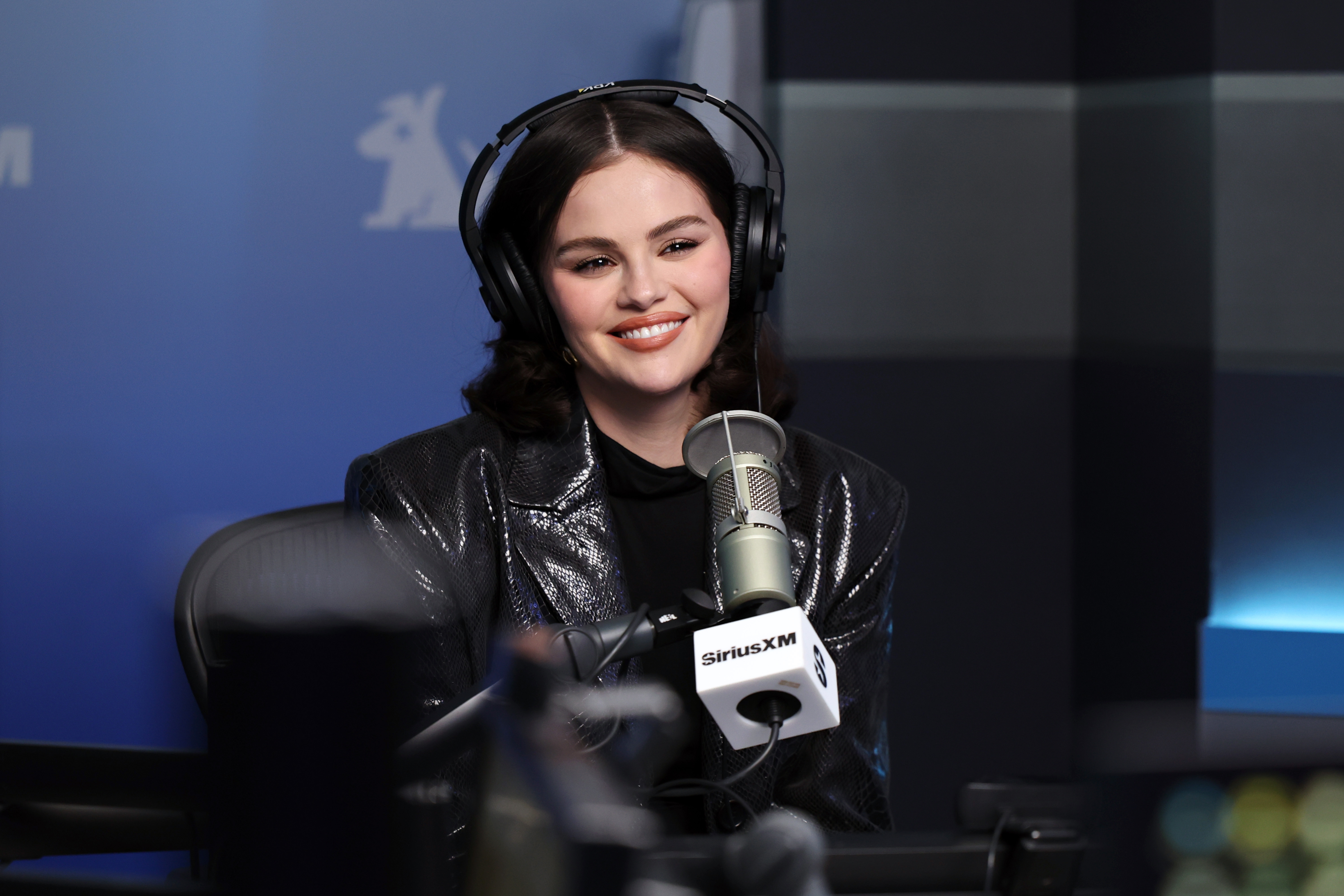 A smiling person sits in a radio studio, wearing headphones and a shiny jacket, with a microphone labeled "SiriusXM" in front of them
