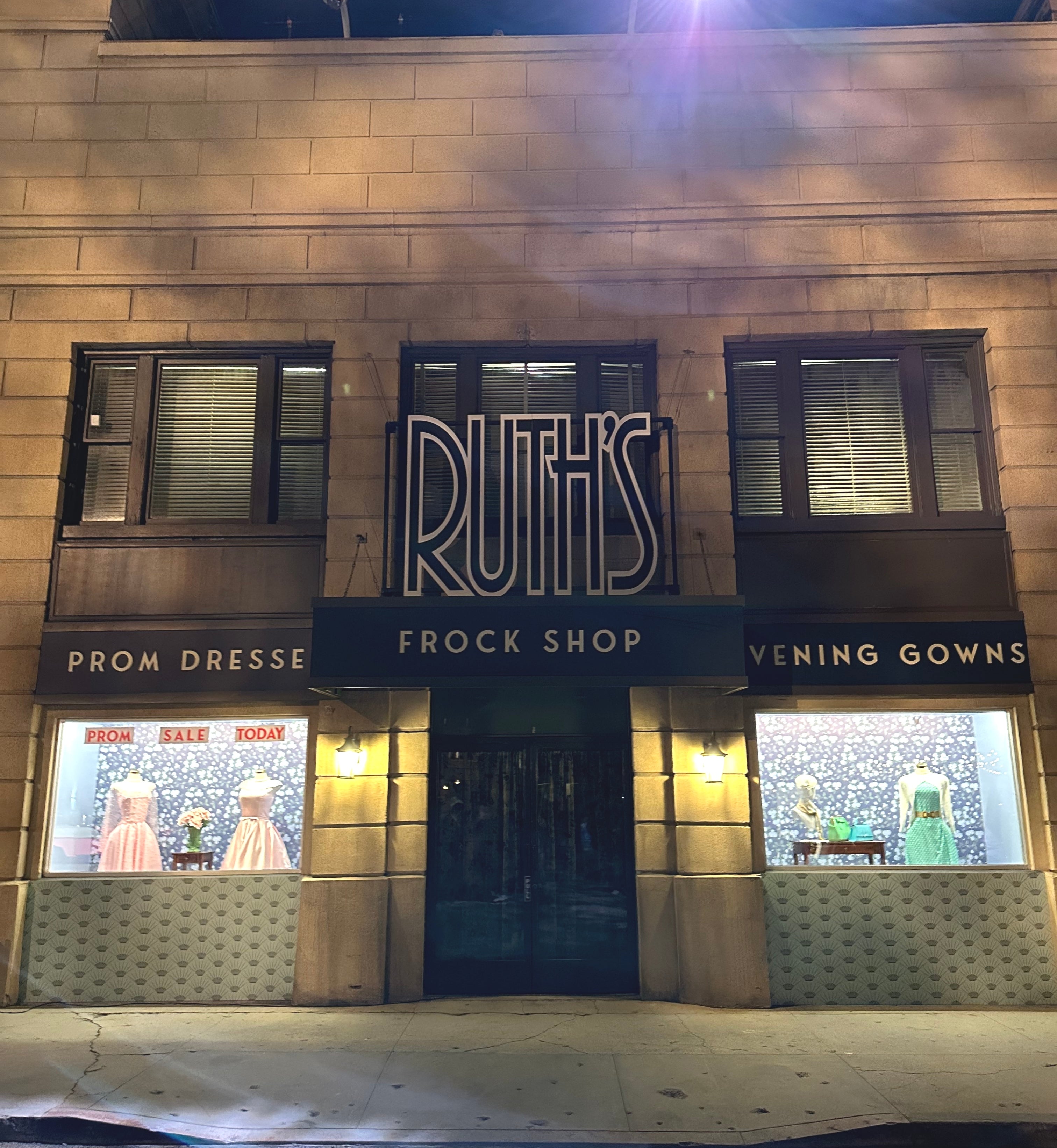 Storefront of Ruth's Frock Shop at night, displaying prom dresses and evening gowns in window displays