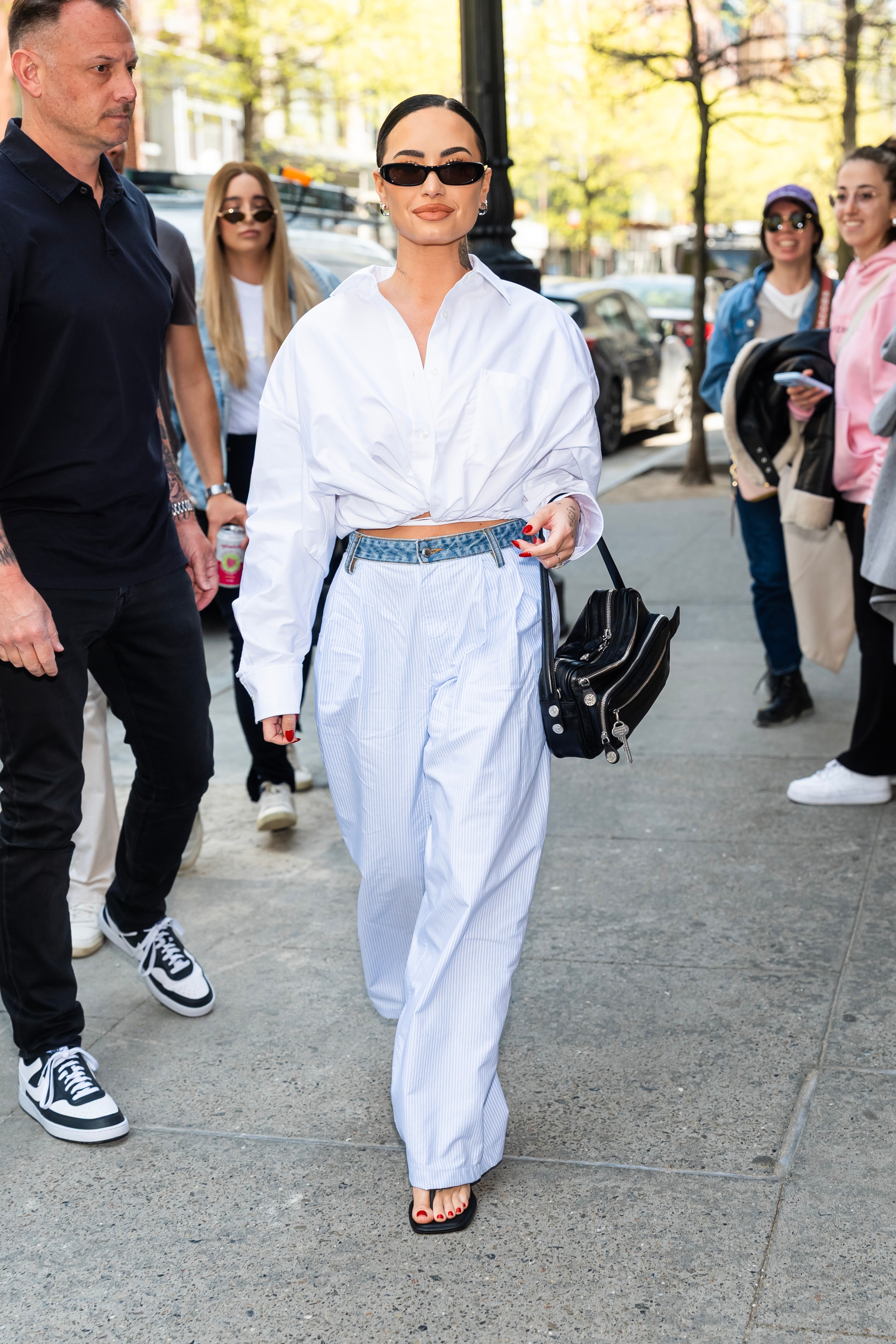 Person in chic wide-leg trousers, cropped shirt, sunglasses, and carrying a black purse, walking confidently on a city street