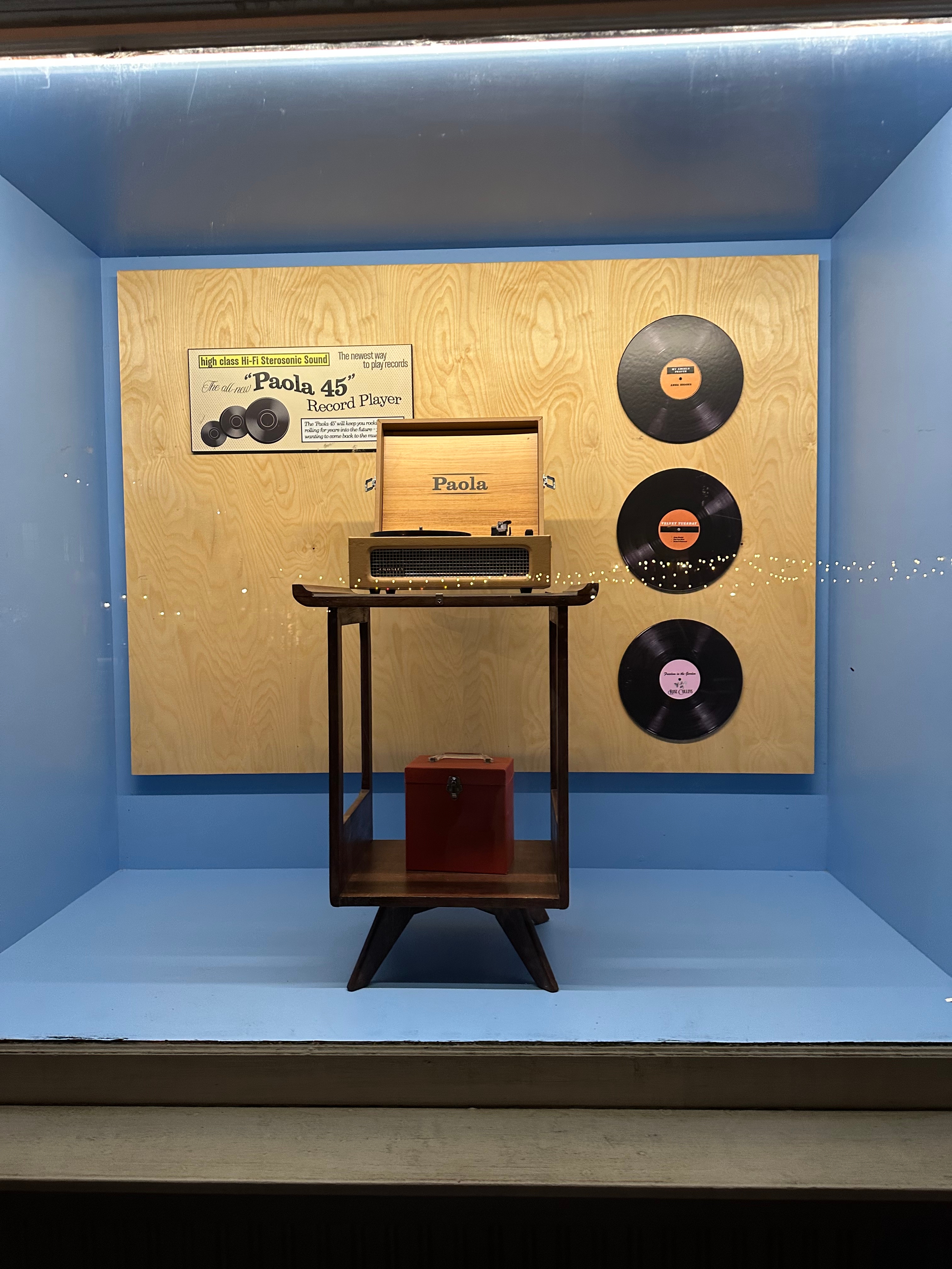 Store window display featuring a vintage Paola 45 record player, three vinyl records on a wooden backdrop, and a small red box on a table