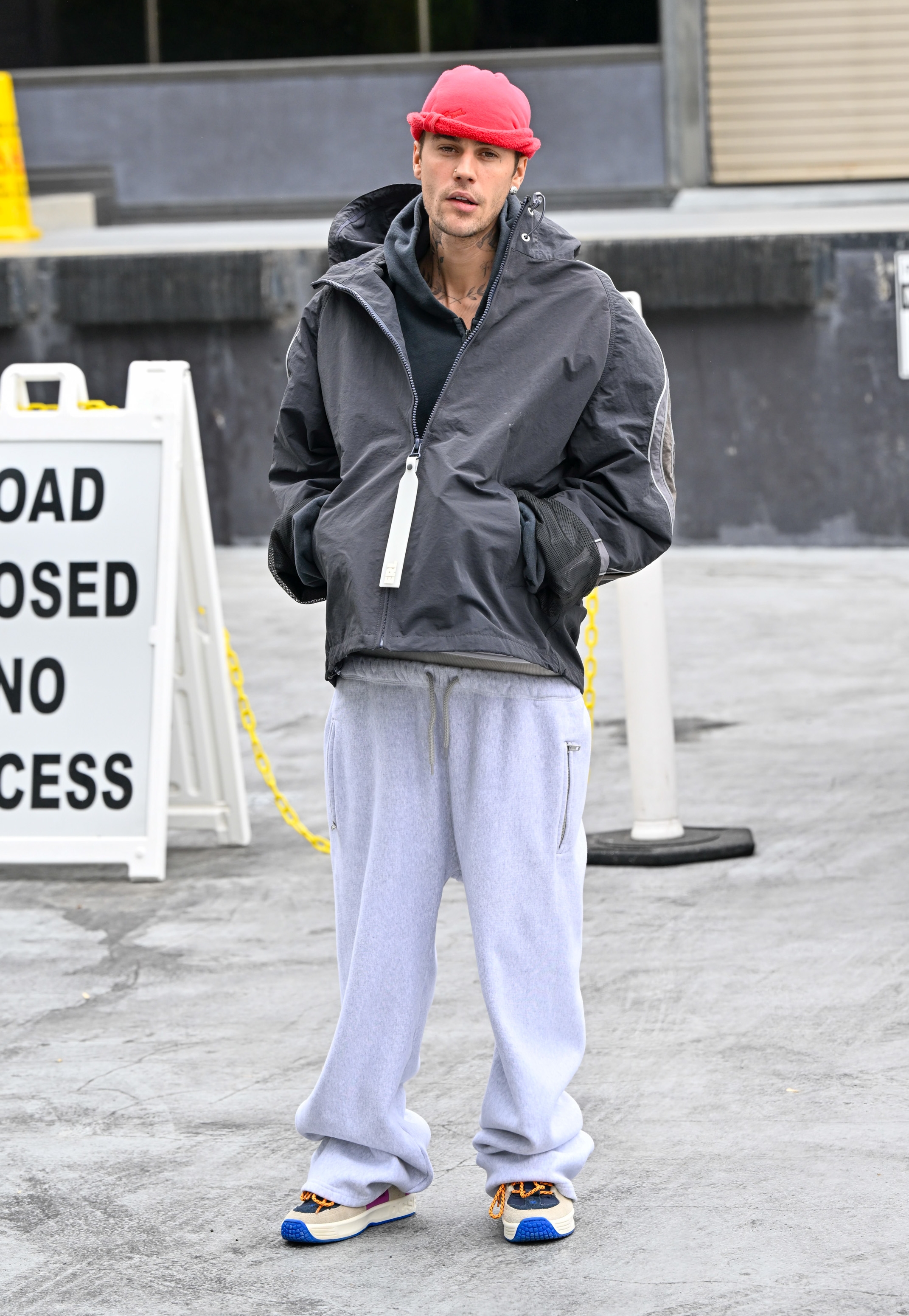 Person in oversized jacket, sweatpants, sneakers, and red beanie standing outside near "Road Closed" sign, conveying a casual, streetwear style