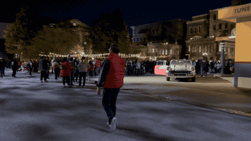 A person in a red vest walks toward a crowd gathered near a classic car under string lights in a street setting at night