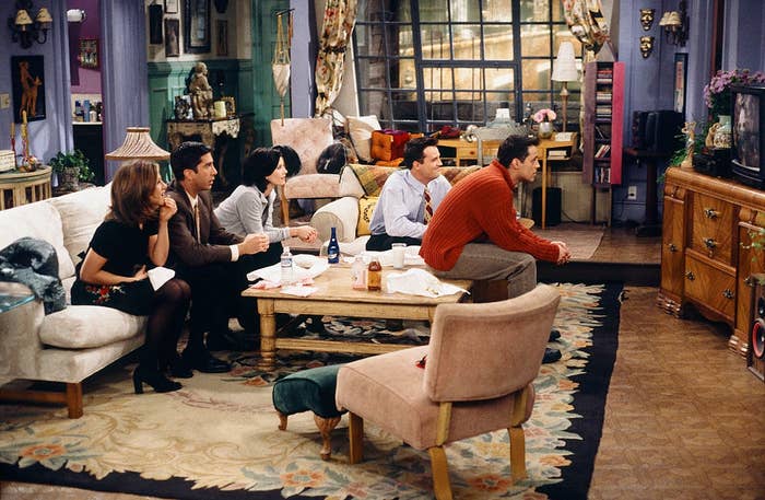 Five people sit on a sofa and chairs, intently watching TV, in a cozy, cluttered living room. The scene is from a popular TV sitcom