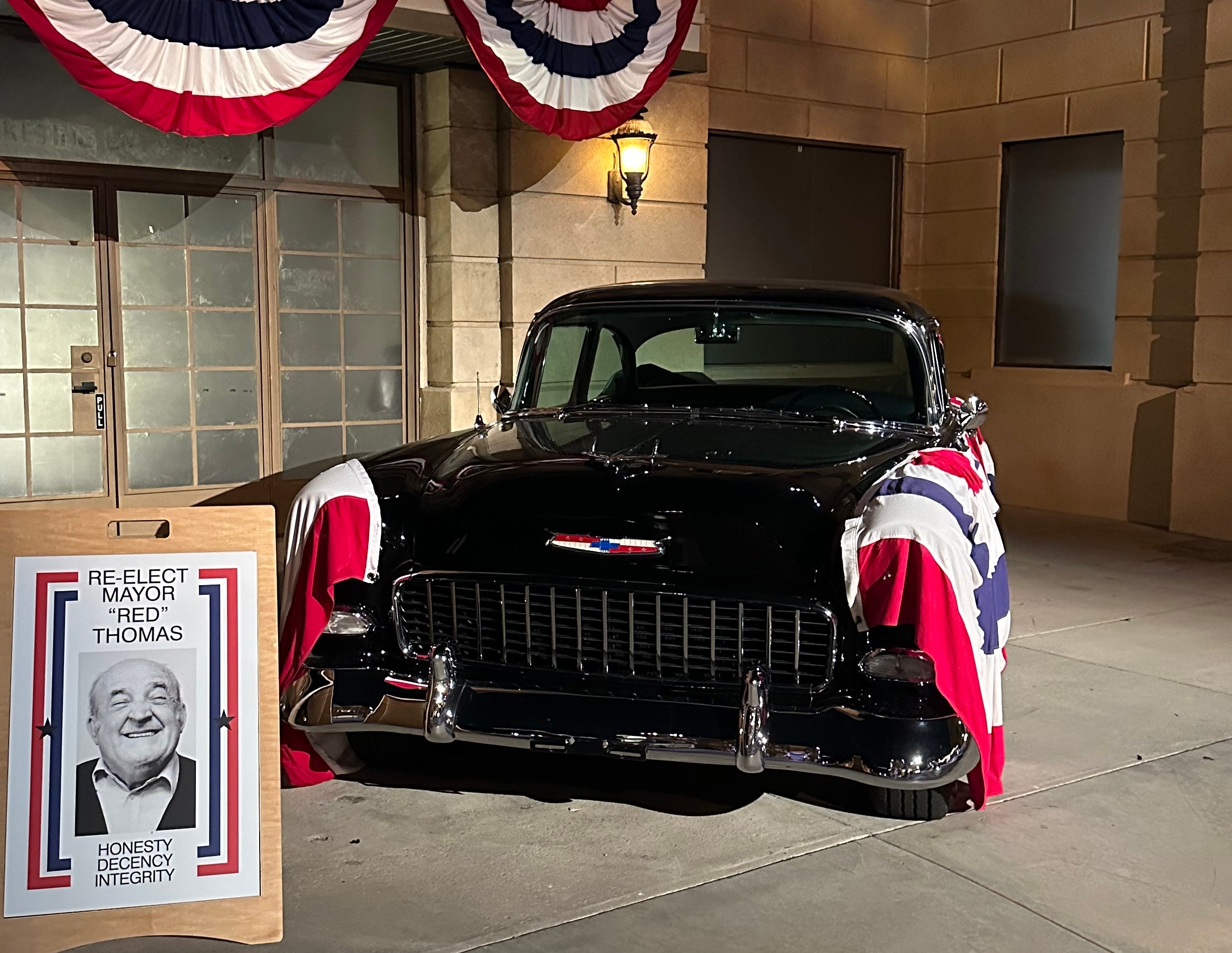Classic car with patriotic banners parked in front of a building. A poster nearby features a person's photo and the text &quot;Re-Elect Thomas.&quot;