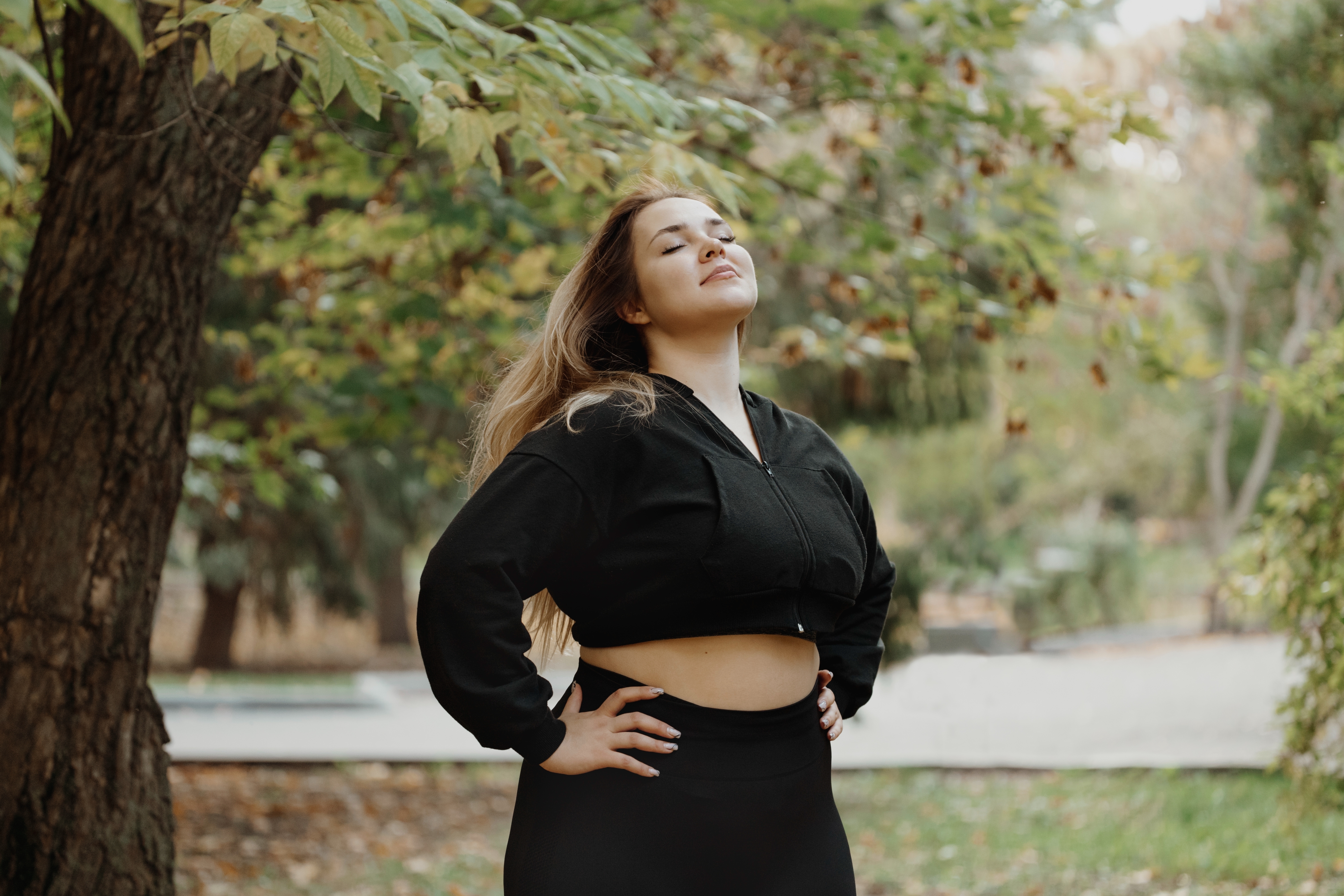 Person in a relaxed outdoor setting, wearing a cropped hoodie and leggings, standing confidently with hands on hips