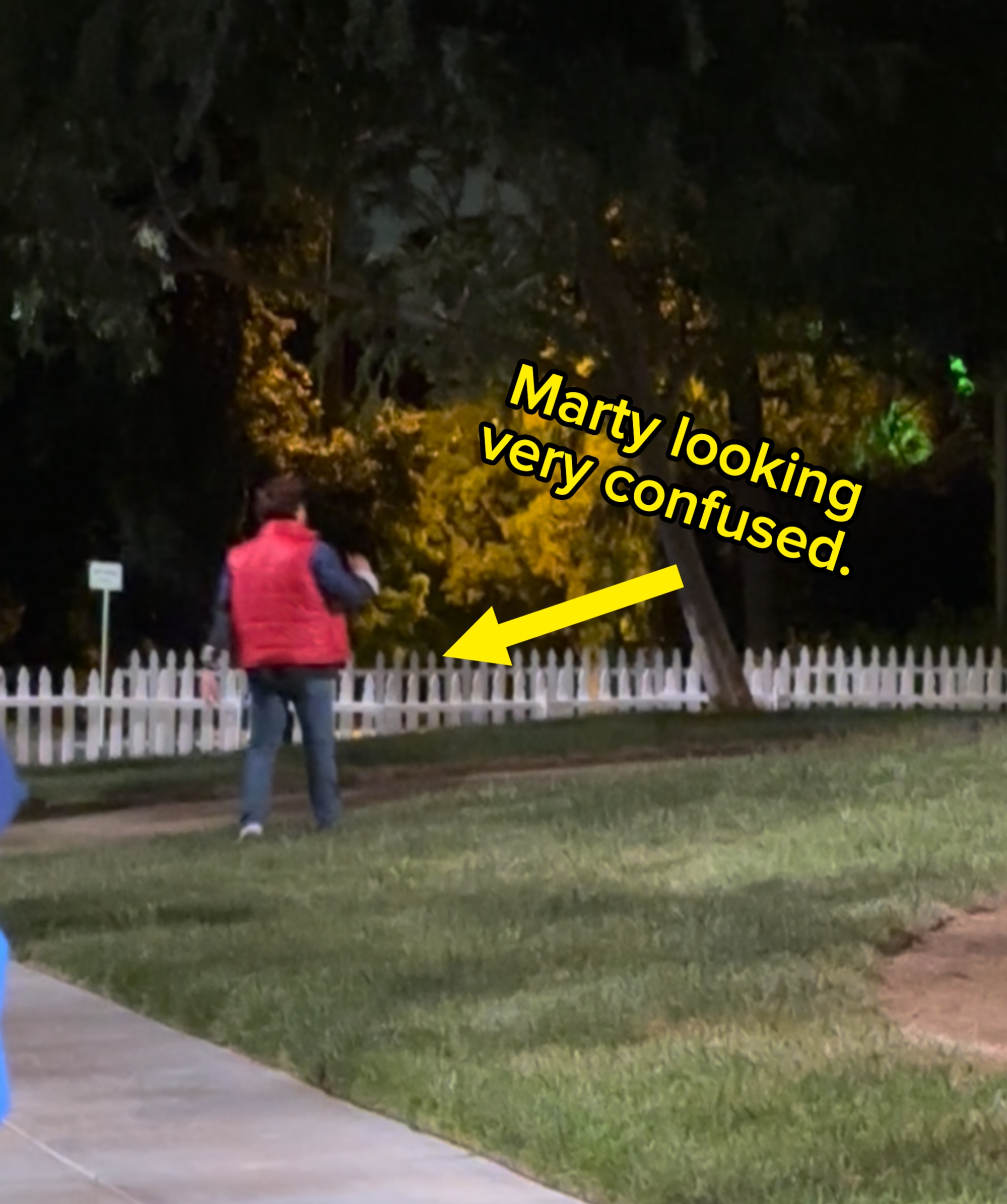 A person in a vest walks on a path near a white picket fence, surrounded by trees at night