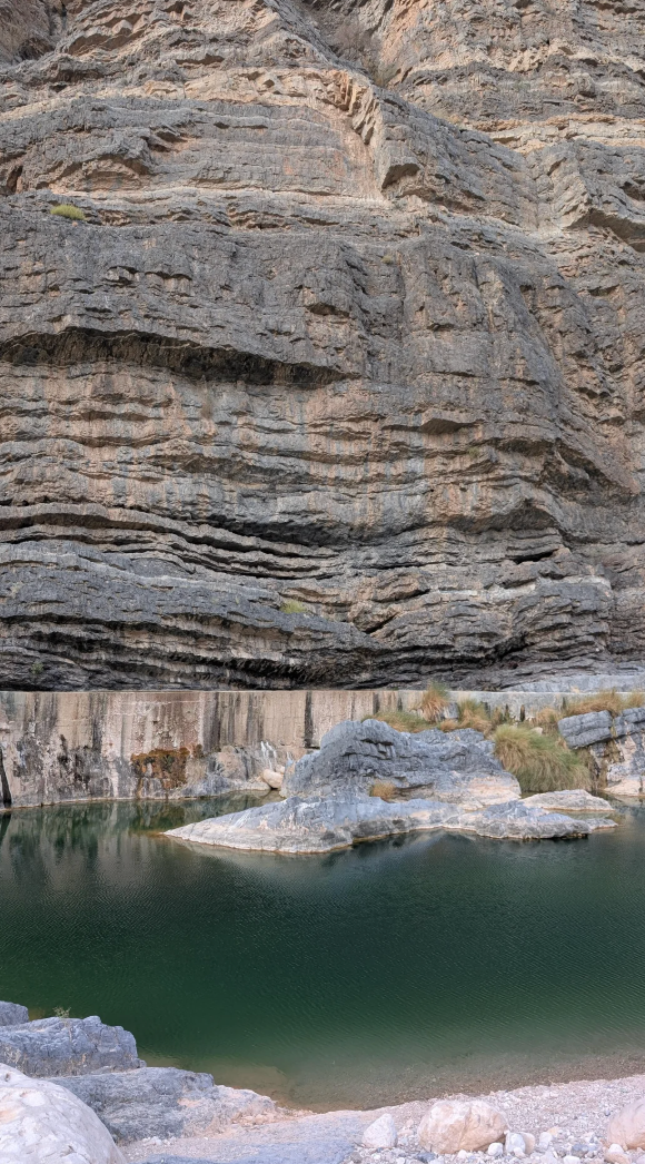 Rocky cliff face with a small, calm pond at its base, surrounded by gravel and sparse vegetation