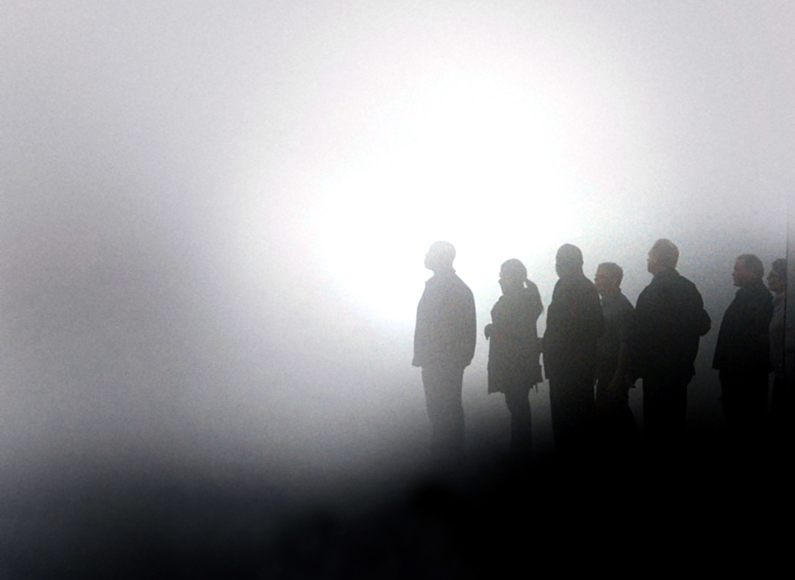 Silhouettes of several people standing in a foggy, indistinct environment, looking towards a bright light