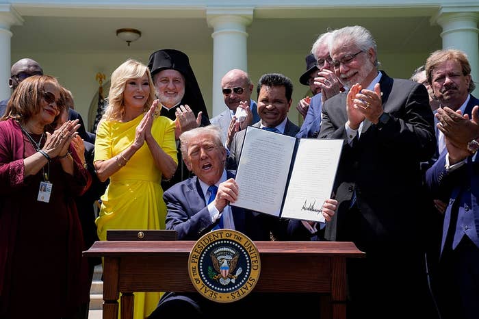 A group of people applauds as a man sitting at a desk holds up a signed document outdoors, with others standing nearby