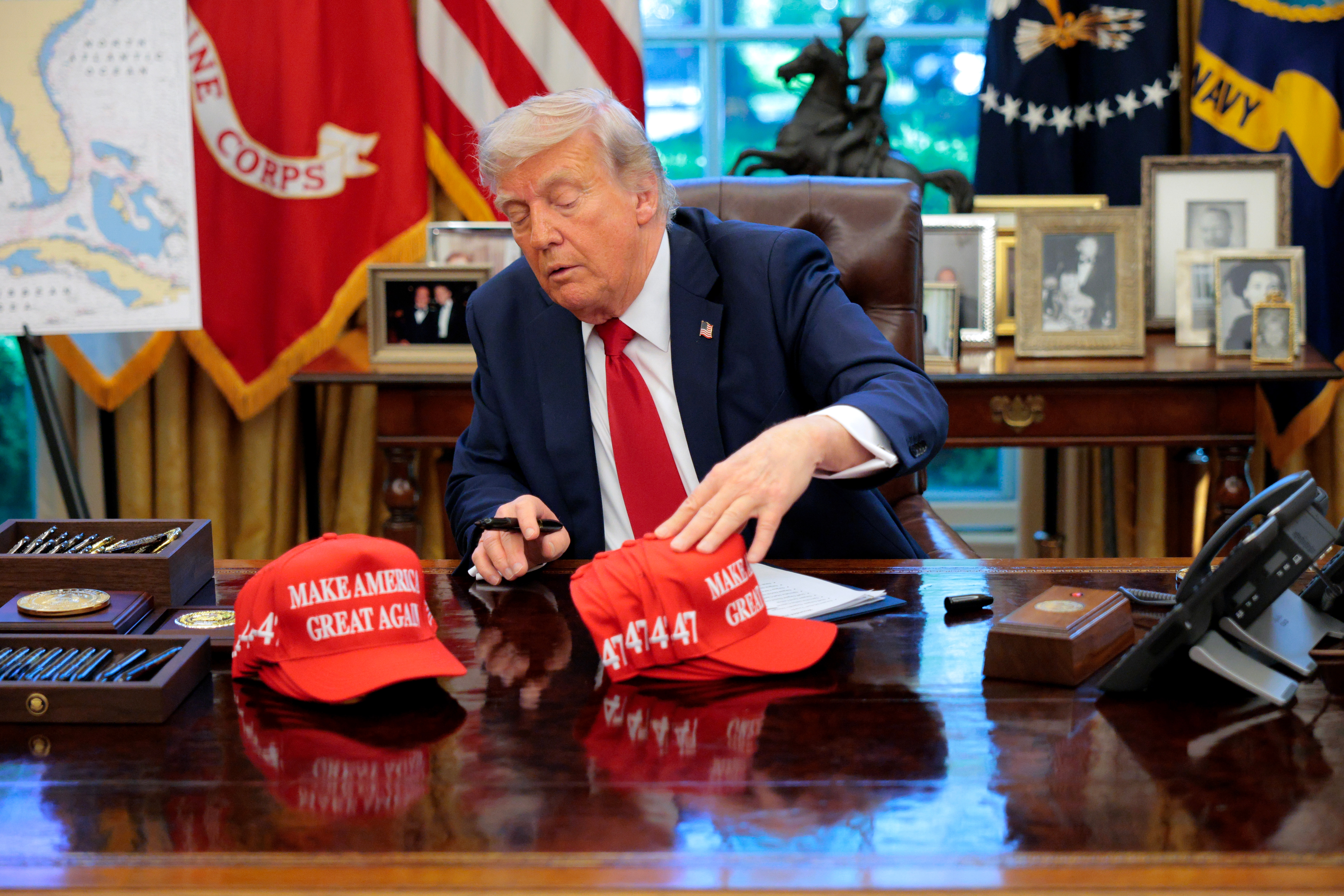 A person in a suit sits at a desk, handling "Make America Great Again" hats in an office setting with flags and framed photos in the background